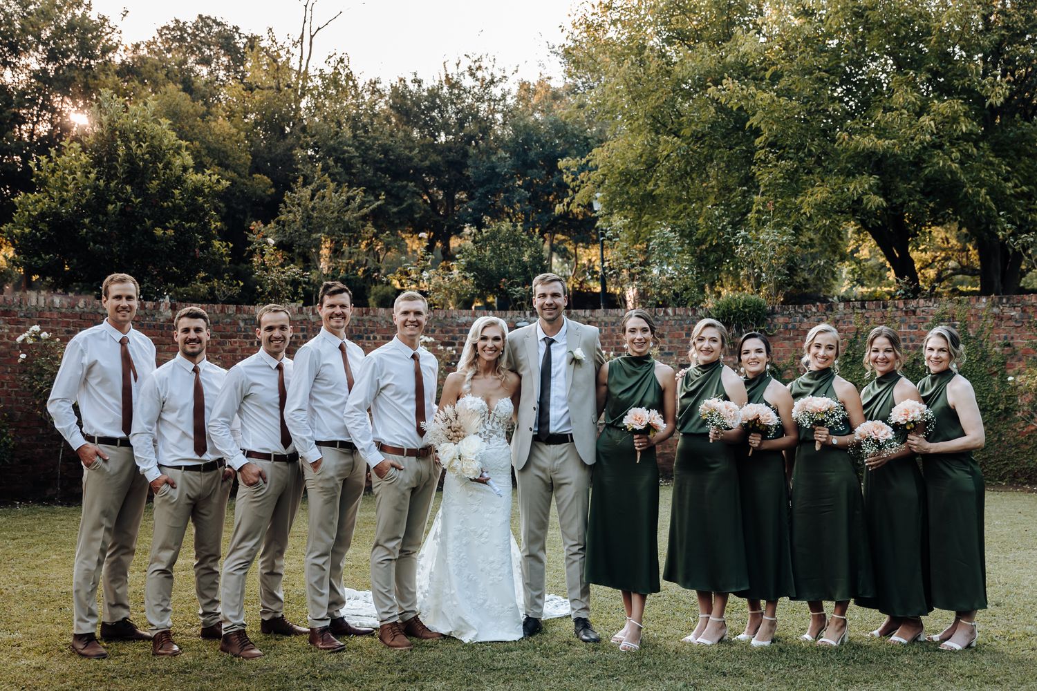 Wedding party posed together on lawn with bridesmaids in dark dresses and groomsmen in light colored attire.