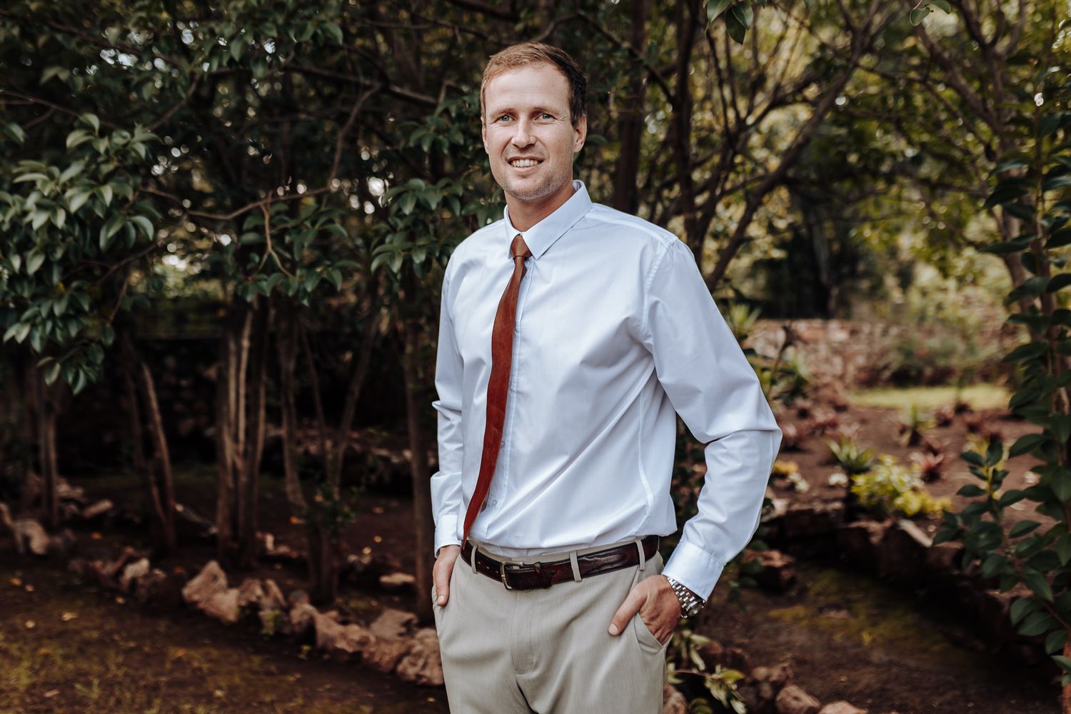 Professional portrait of man in business attire standing against natural wooded backdrop.
