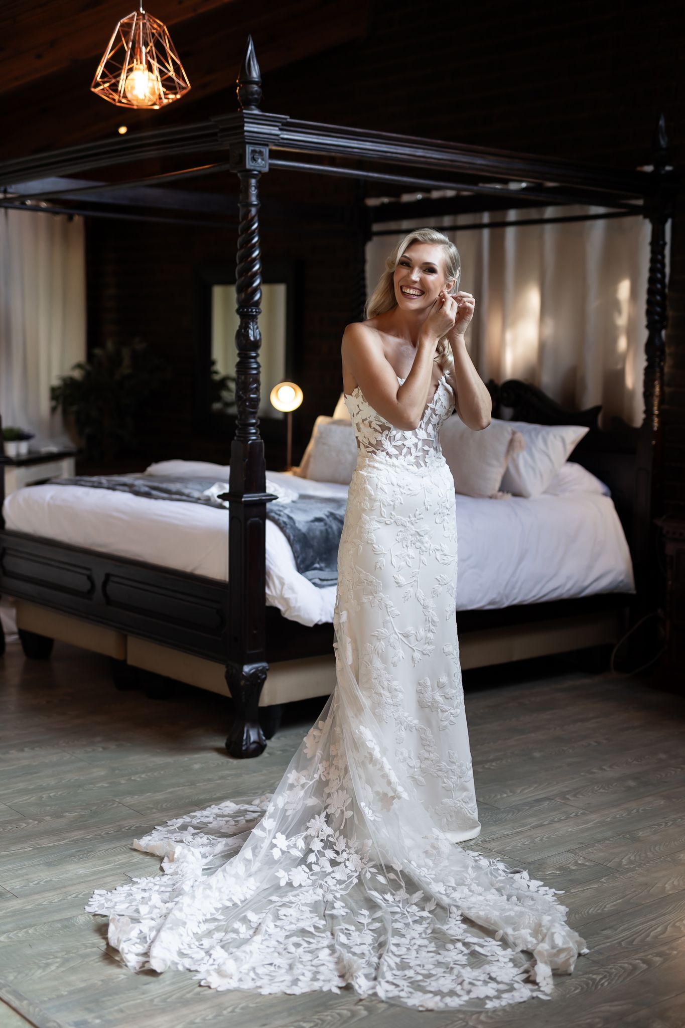 Bride wearing fitted lace wedding dress smiles while standing beside four-poster bed.