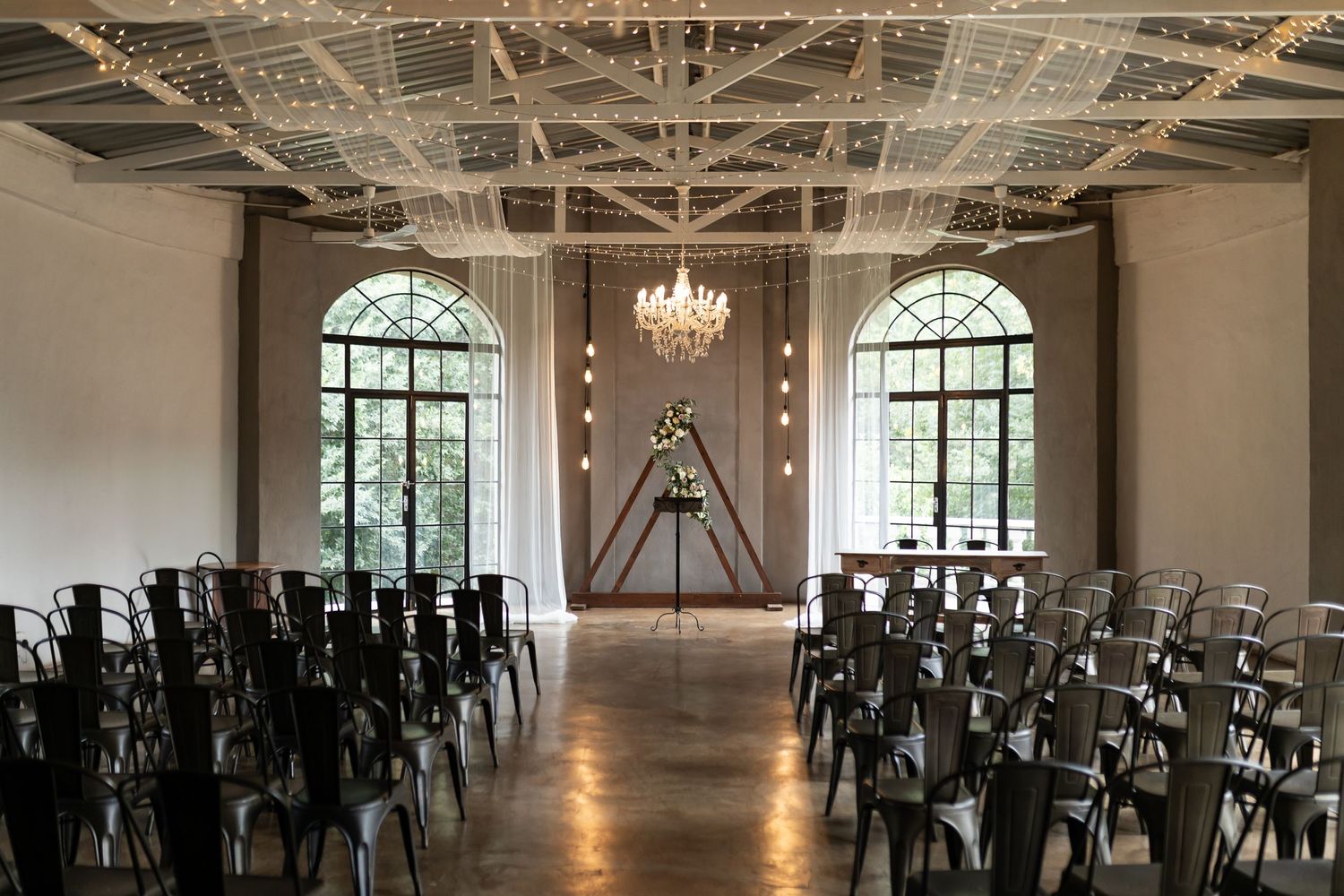 A modern wedding ceremony space with arched windows, black chairs and crystal chandeliers.