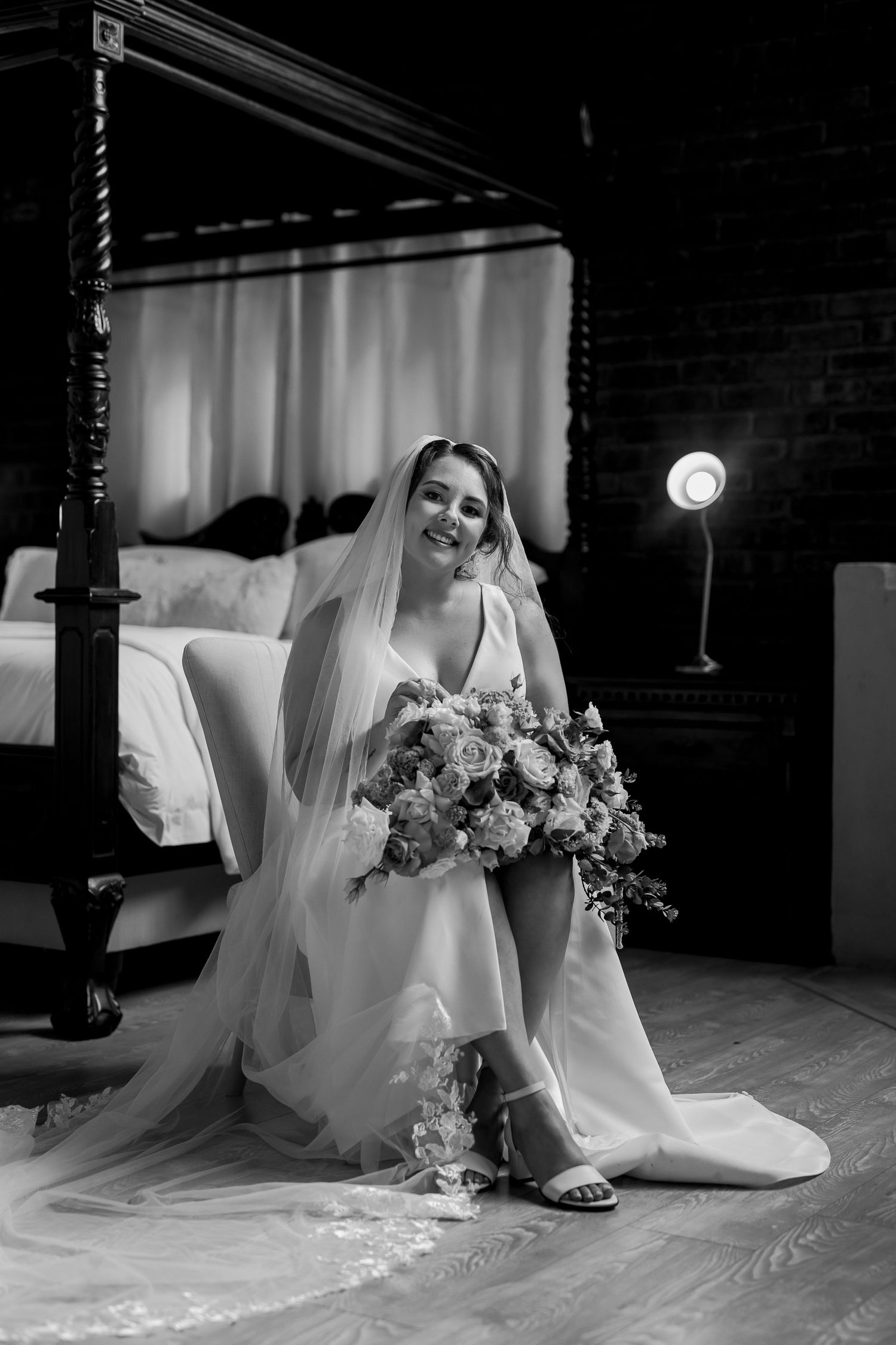 Black and white photo of a bride seated with flowers near a canopy bed and glowing lamp.