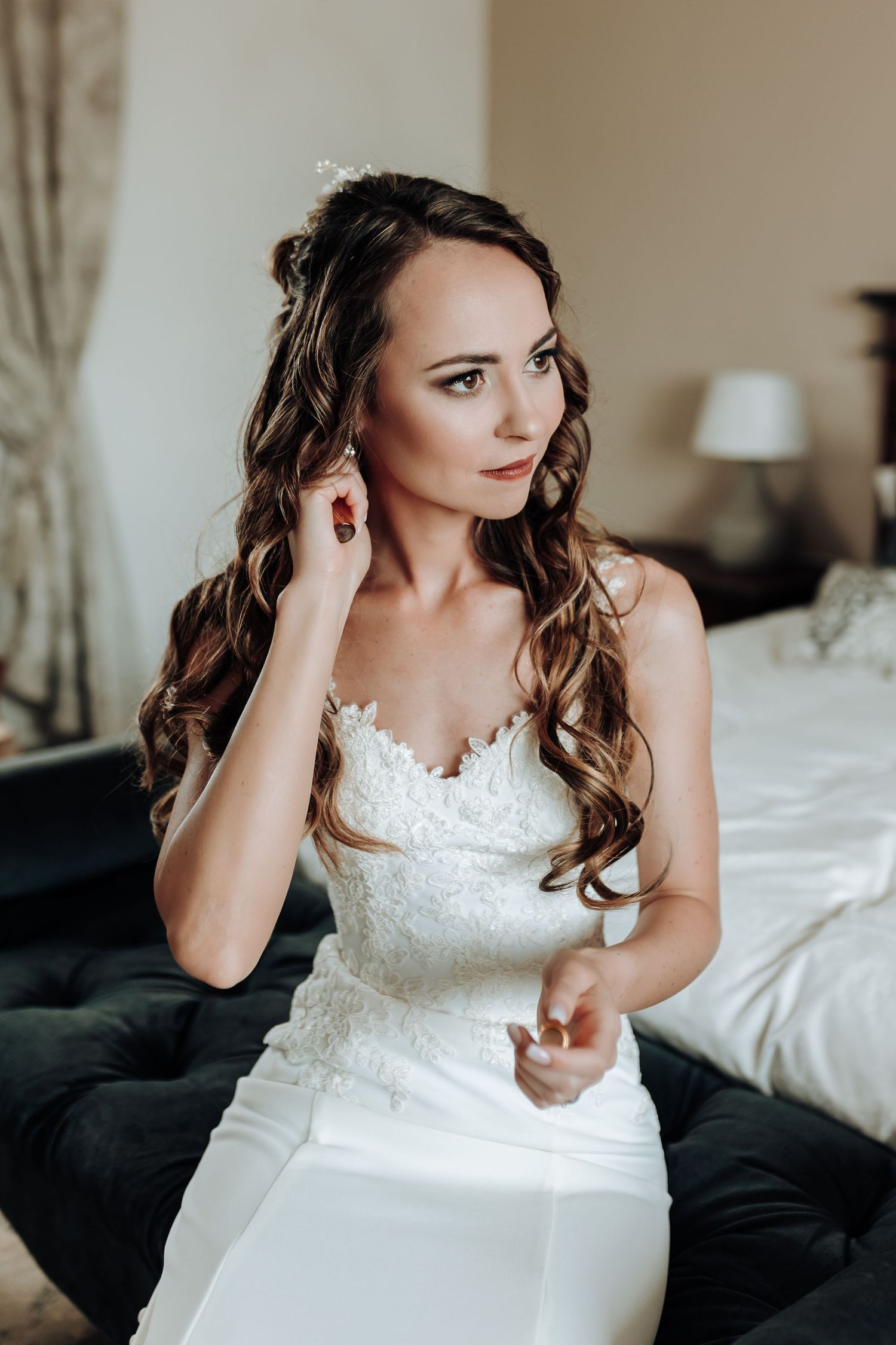 Elegant bride in white lace gown sitting on dark couch with romantic hairstyle.