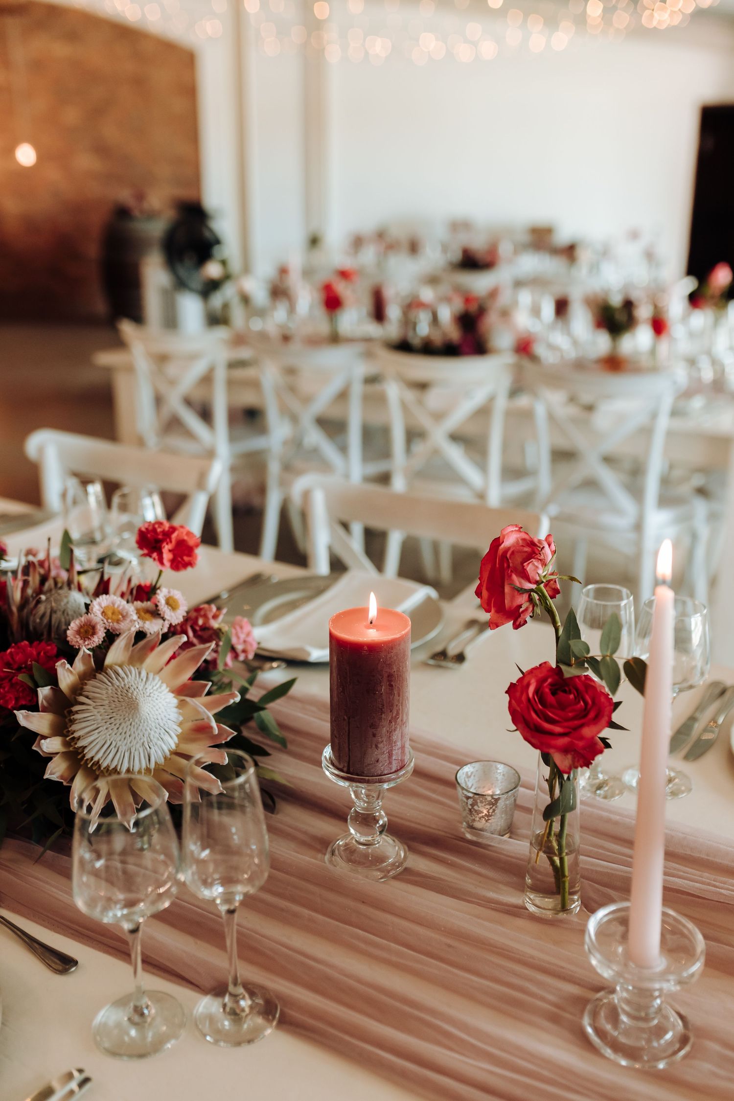 Wedding table setting with red roses, protea flowers, and burgundy candles in an elegant reception hall.