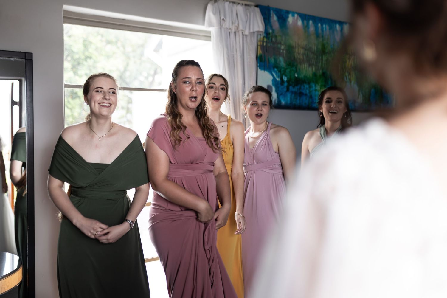 Bridesmaids in different colored dresses react with joy while looking at someone off camera.