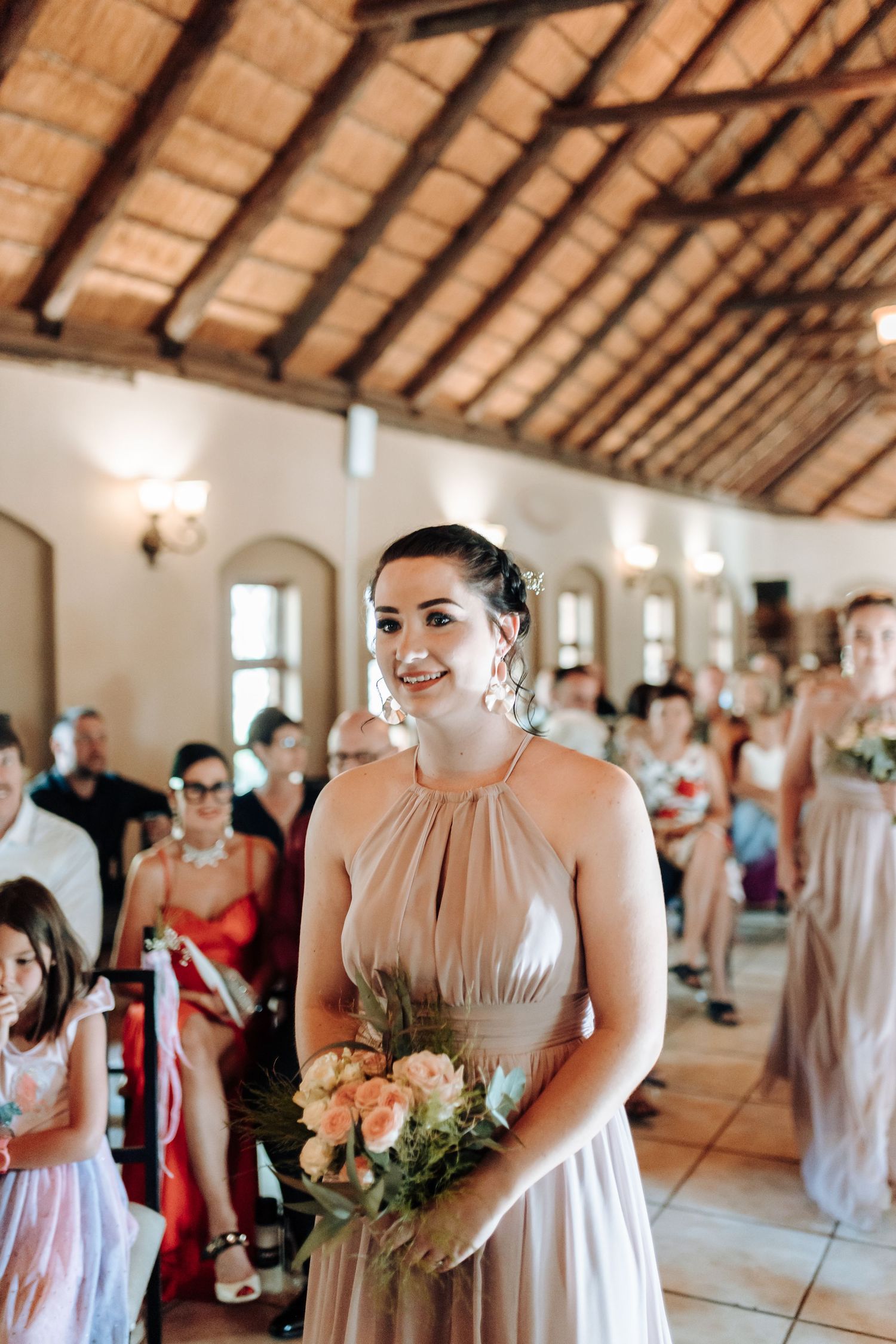 A bridesmaid smiles while holding a peach bouquet in an elegant venue with exposed wooden beams.
