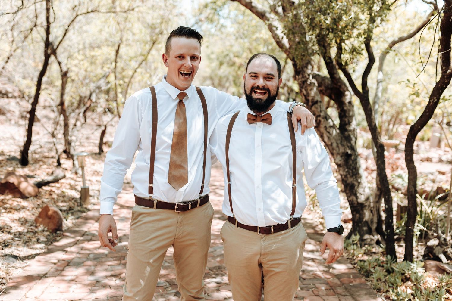 Two groomsmen in matching khaki pants and dress shirts pose together on a wooded path at a wedding.