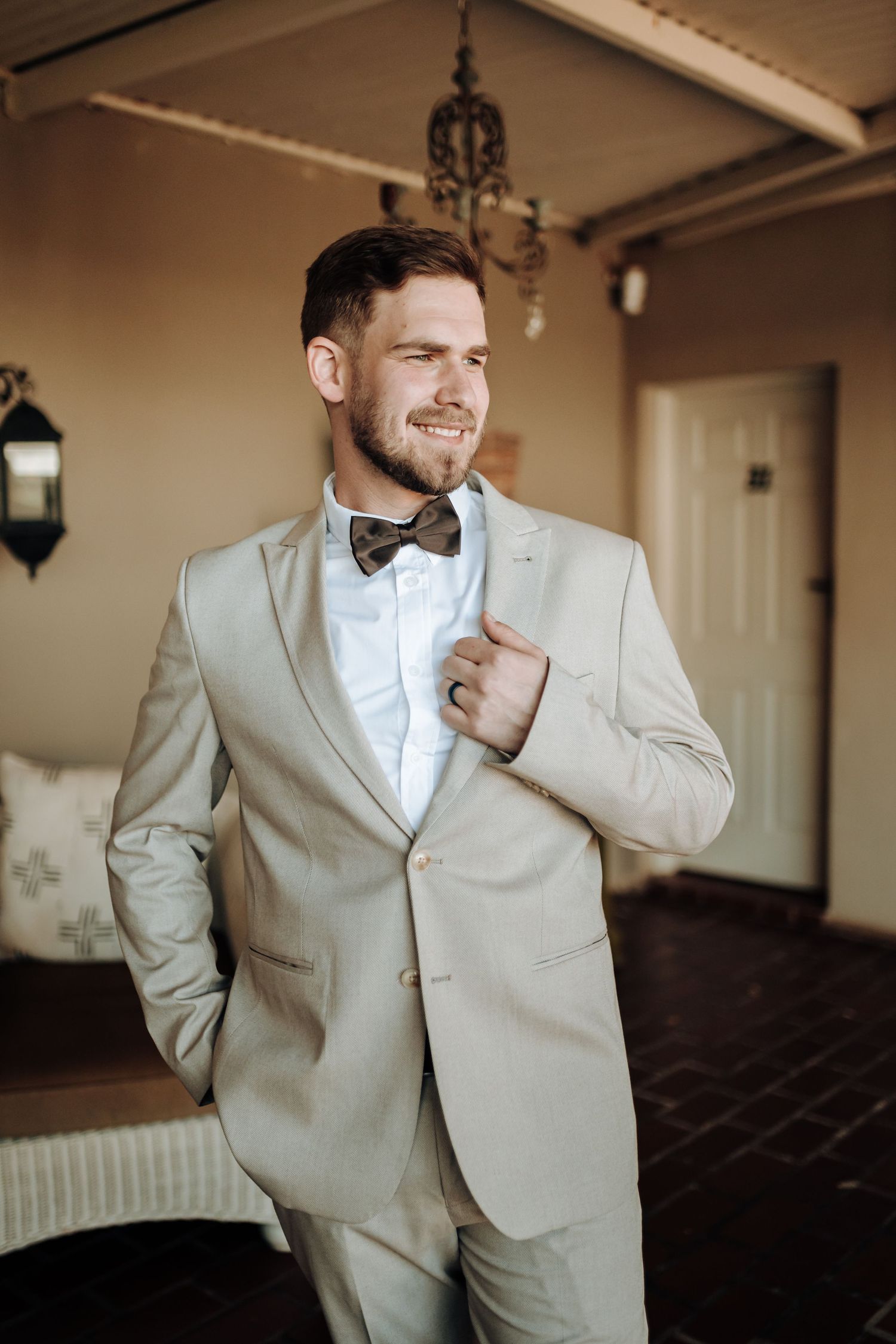 A groom in a light beige suit and bowtie smiles while adjusting his jacket in an elegant indoor setting.
