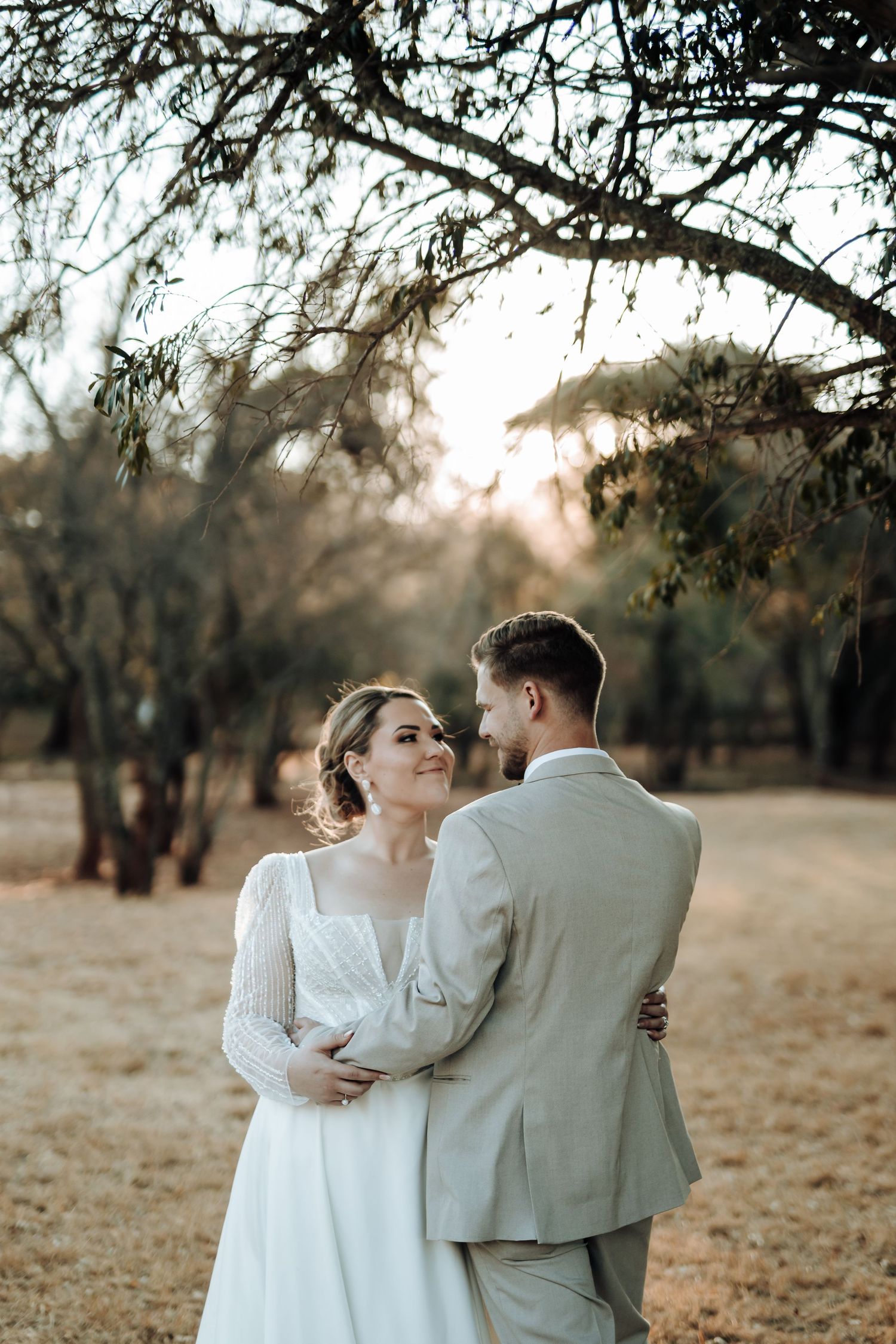 Romantic wedding couple embraces at sunset in a rustic outdoor setting with trees and golden light.