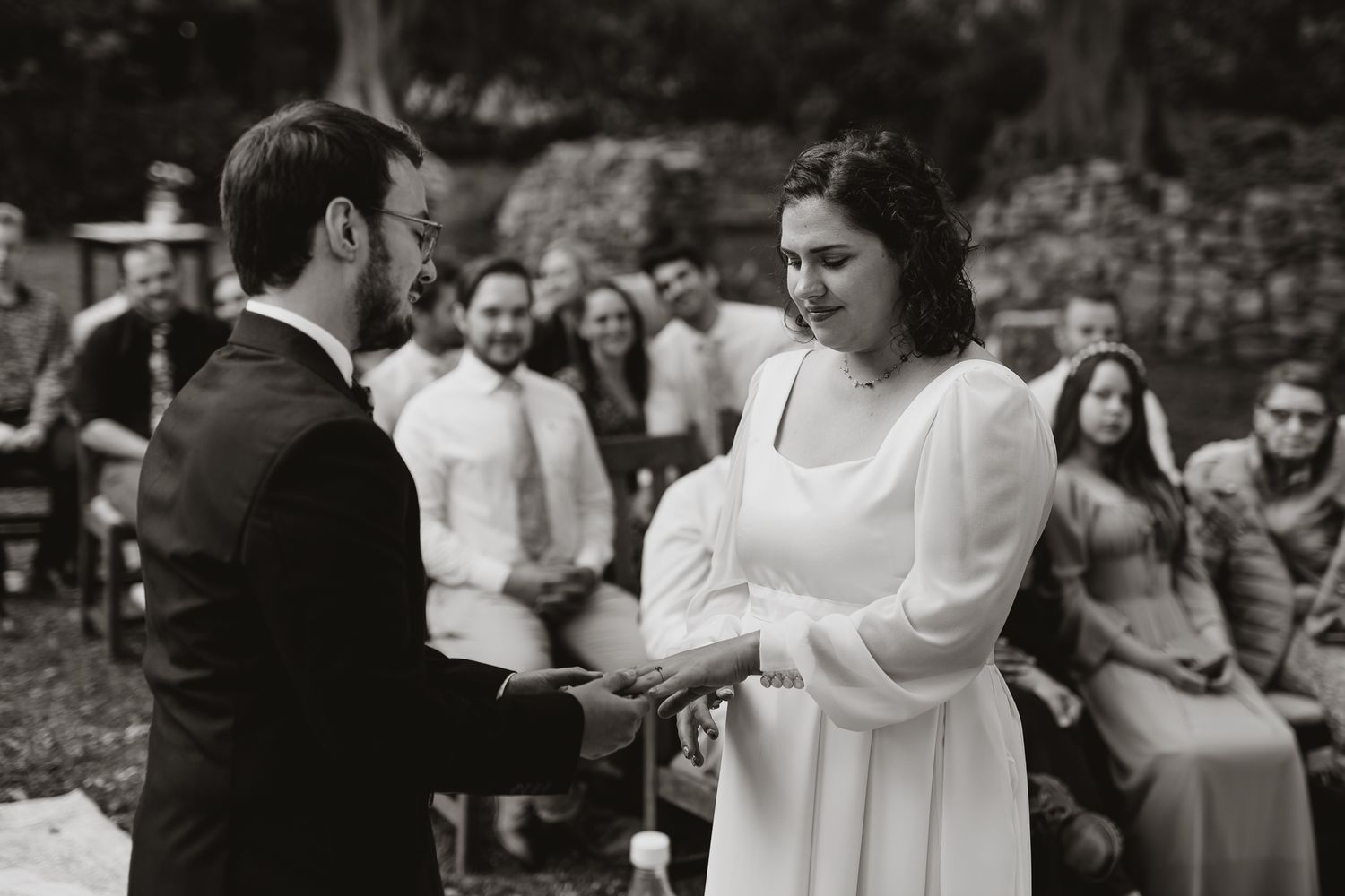 A black and white photograph captures an intimate moment during a wedding ceremony in a garden setting.