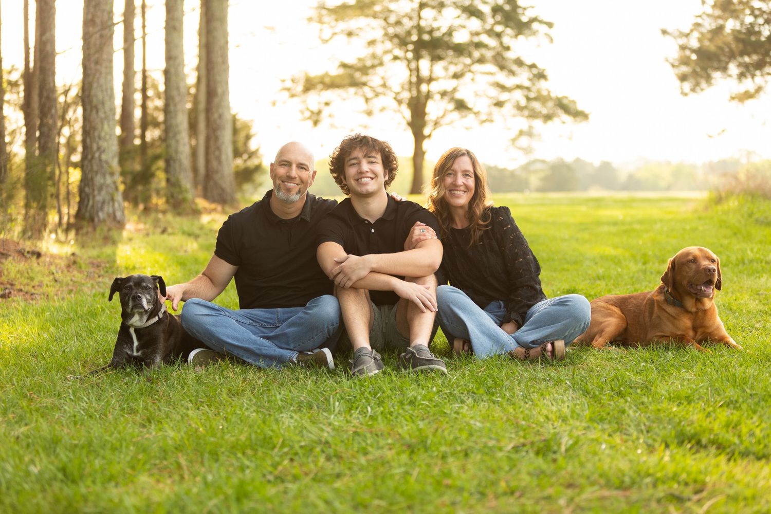 A family sits together on grass near tall pine trees with their two dogs during an outdoor photo session.