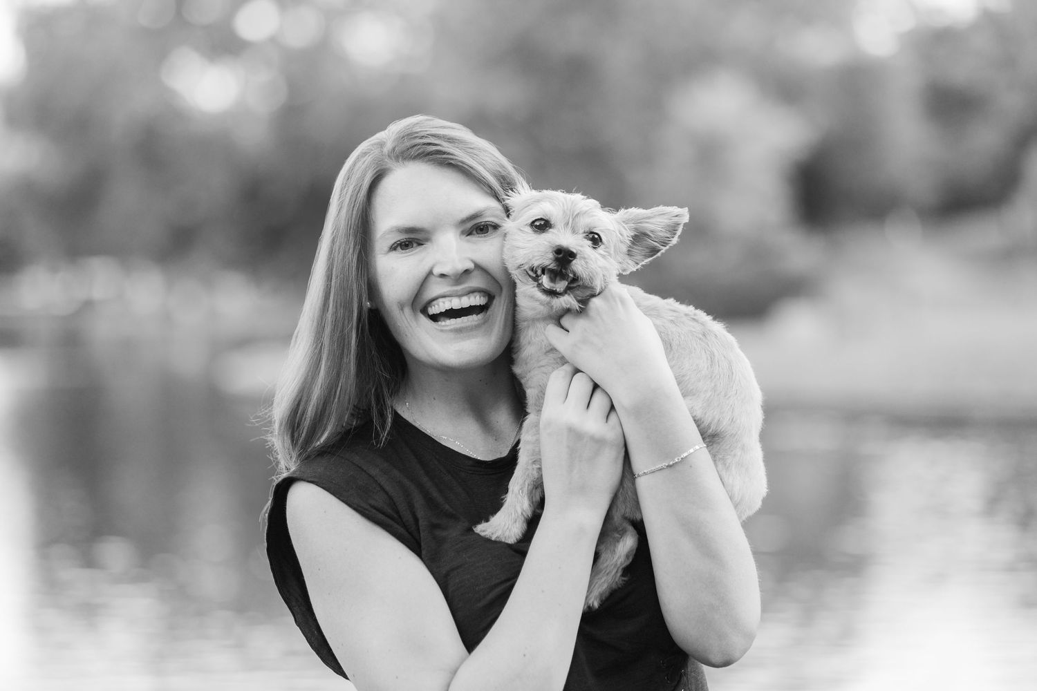 Black and white portrait of someone holding and embracing a small smiling white Chihuahua puppy outdoors.