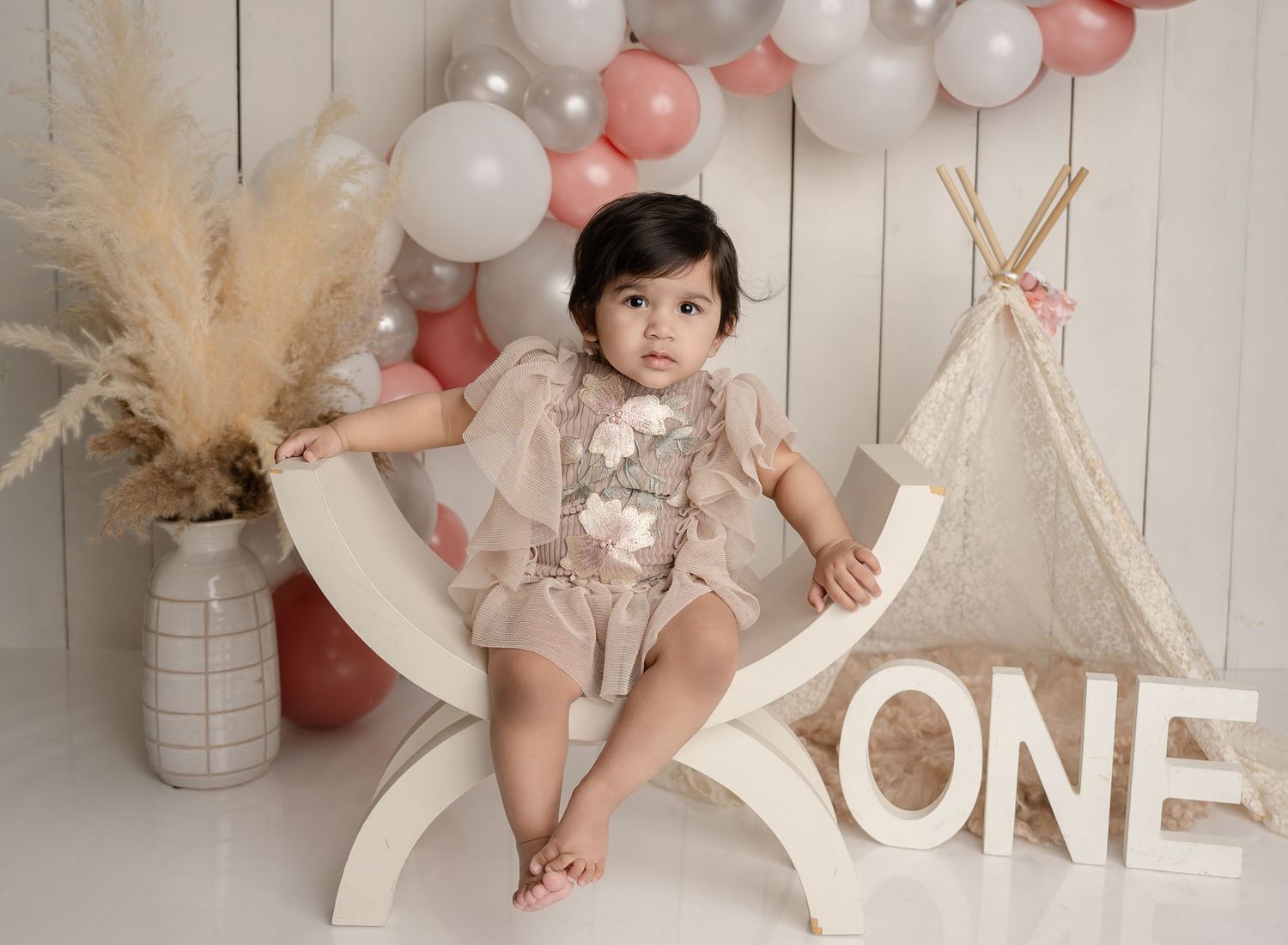 First birthday photo setup with white and pink balloons, teepee tent, pampas grass and 'ONE' sign against wooden backdrop.
