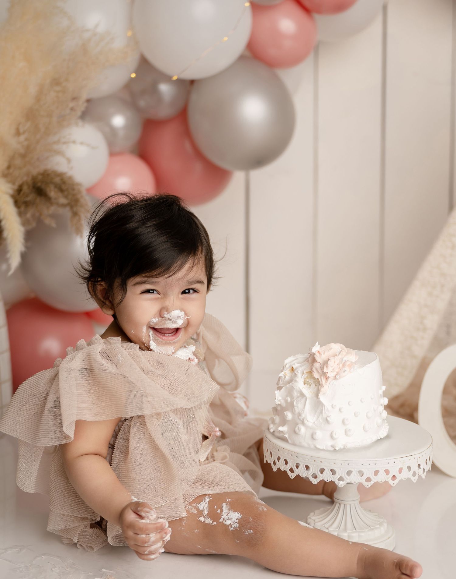 A toddler in a cream-colored dress sits beside a decorated white cake with balloon decorations in pink, silver and white.