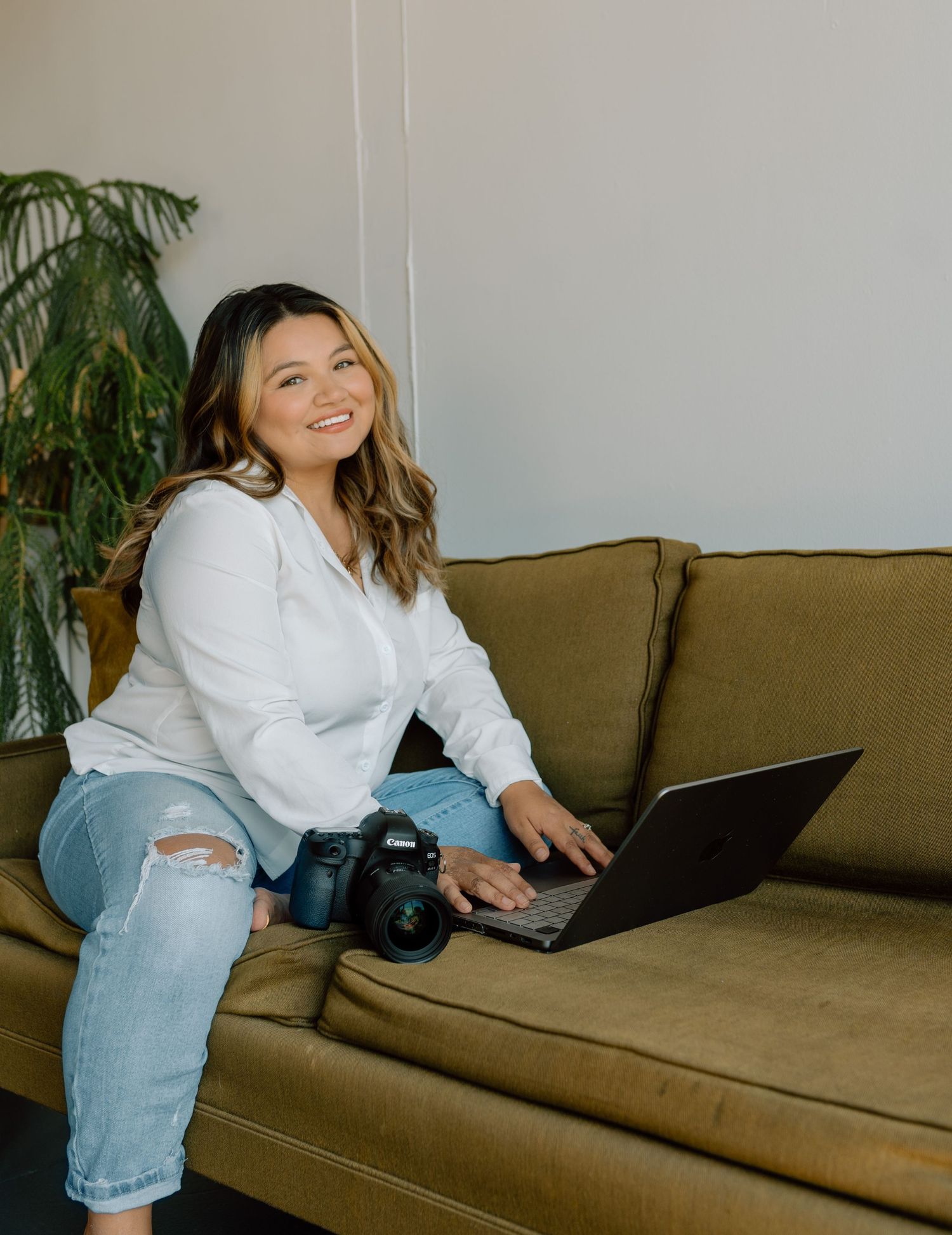 Someone sits on a brown couch working on a laptop with a camera nearby and houseplant in background.