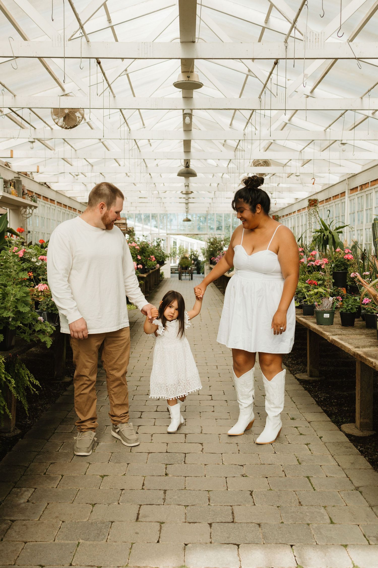 Family in coordinating white outfits walking together in greenhouse filled with flowers and plants.