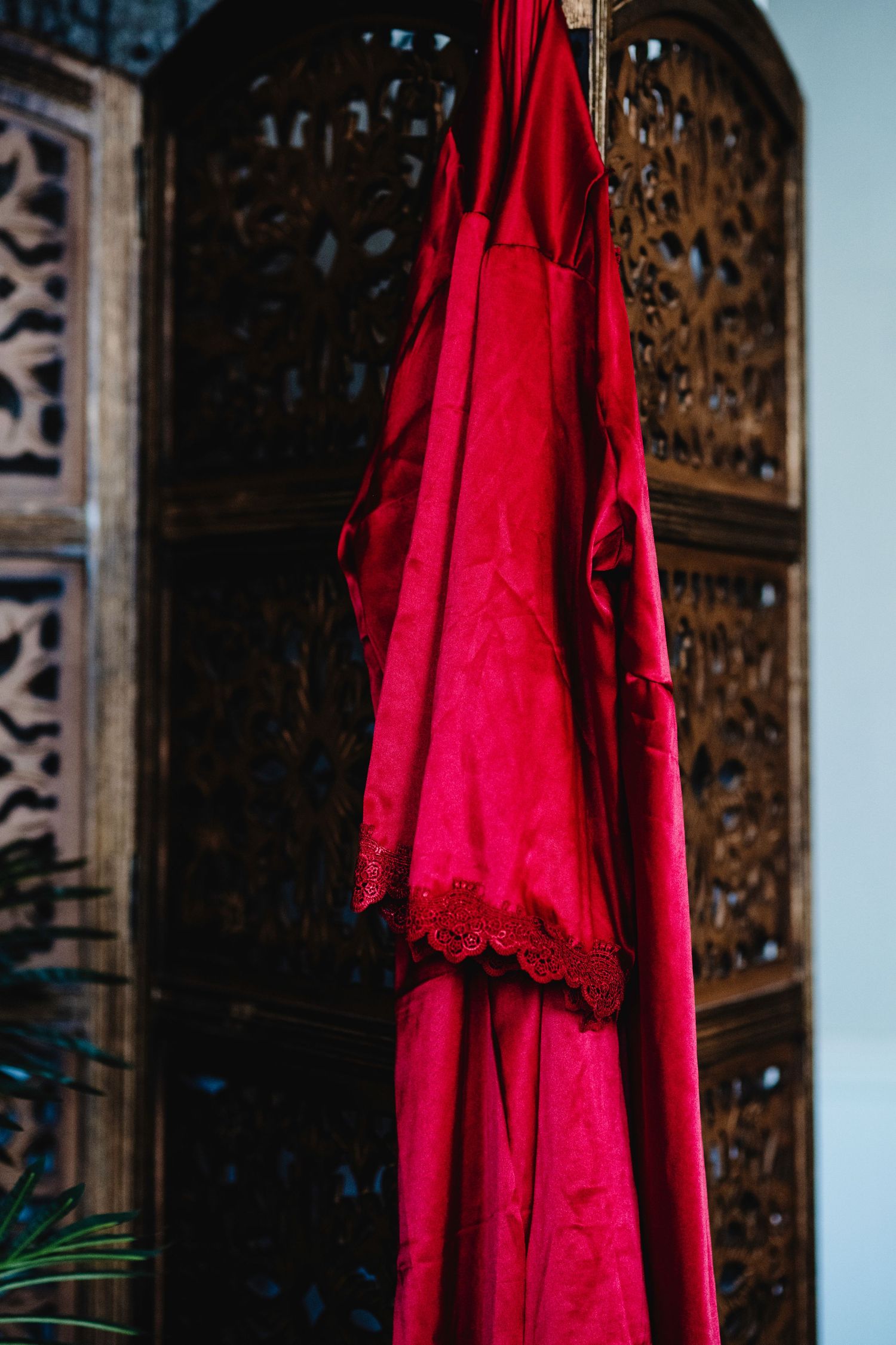 A bright red robe or dress with lace trim hangs against a dark wooden decorative screen with intricate patterns in a dramatic boudoir studio