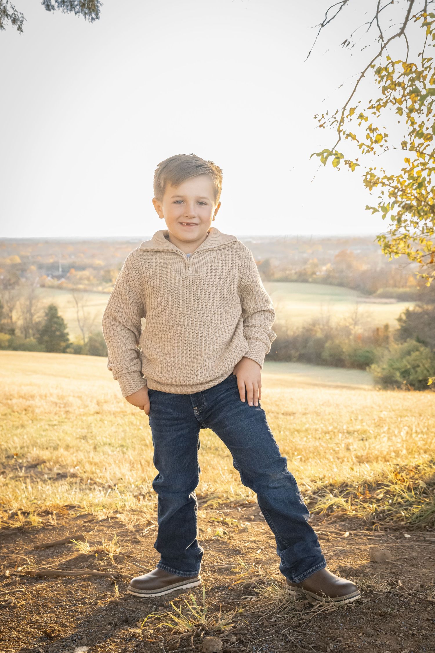 Young child in beige sweater and jeans poses on hilltop during golden autumn sunset with countryside view in background.