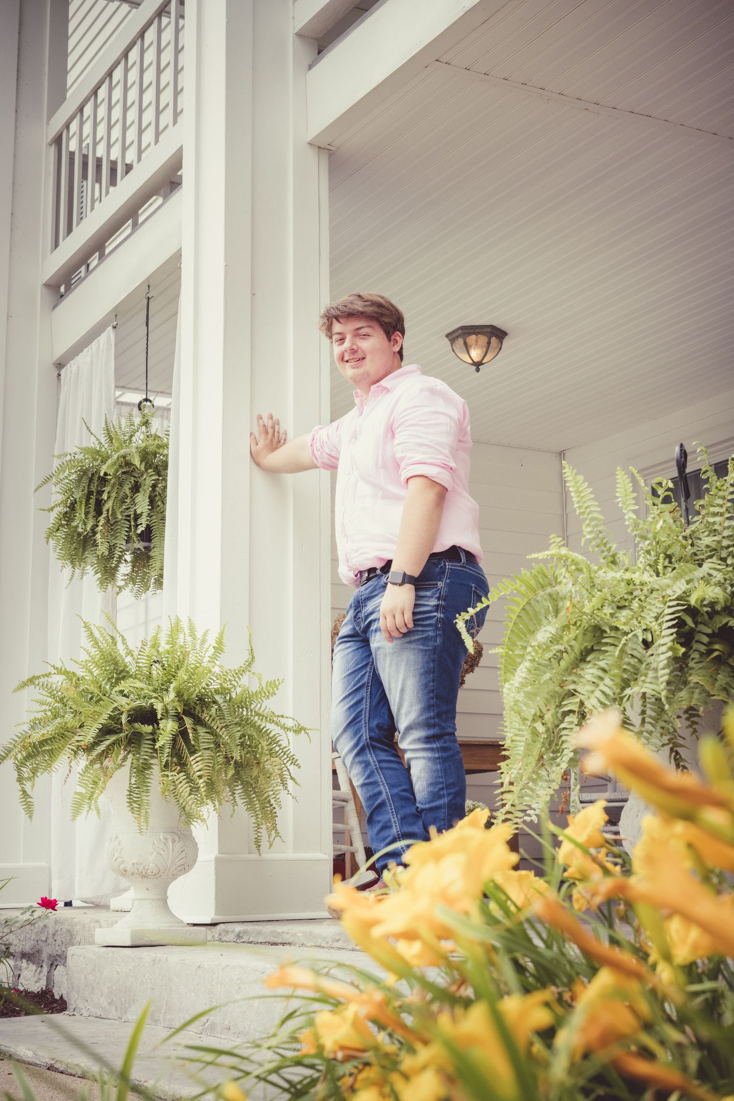 A guy in a pink shirt and jeans leans against a Southern-style porch decorated with hanging ferns and yellow lilies in this Senior Portrait.