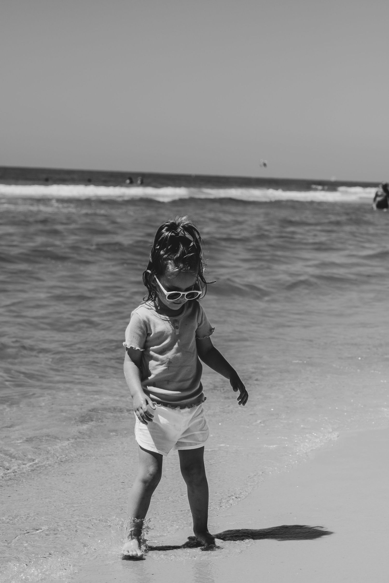 Black and white photo of a small child standing on a beach near ocean waves wearing summer clothing and sunglasses.