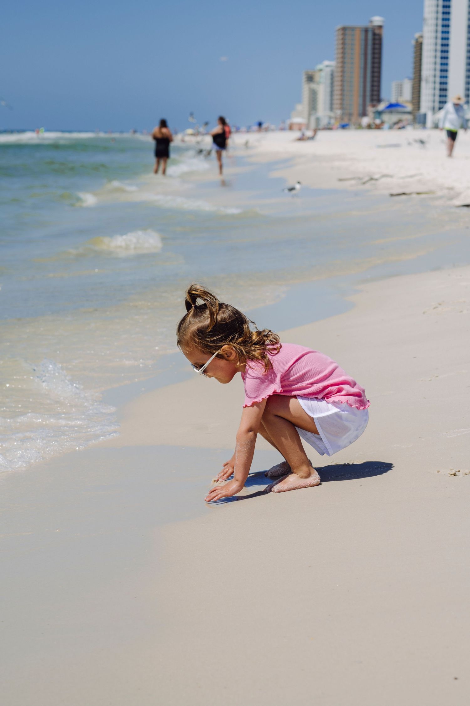 Young child in pink top bends down to explore the sandy beach with high-rise buildings visible along the shoreline.