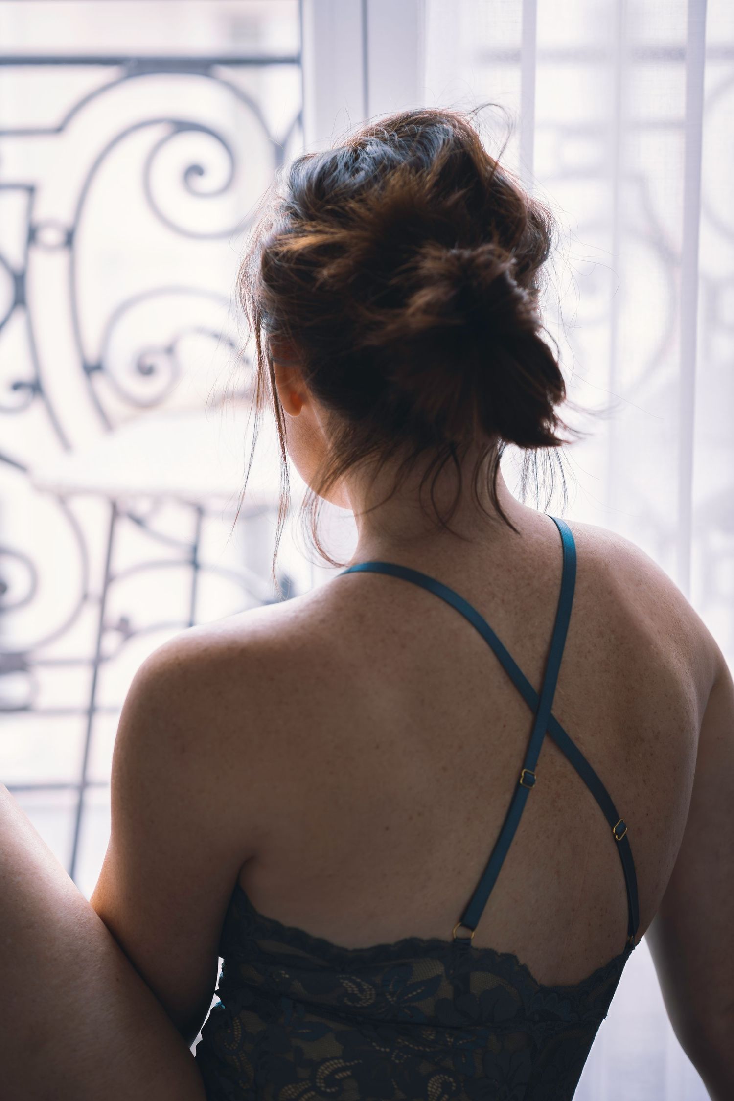 French Riviera Boudoir: Back view of a woman in a green lace camisole looking through an elegant window with ornate ironwork.