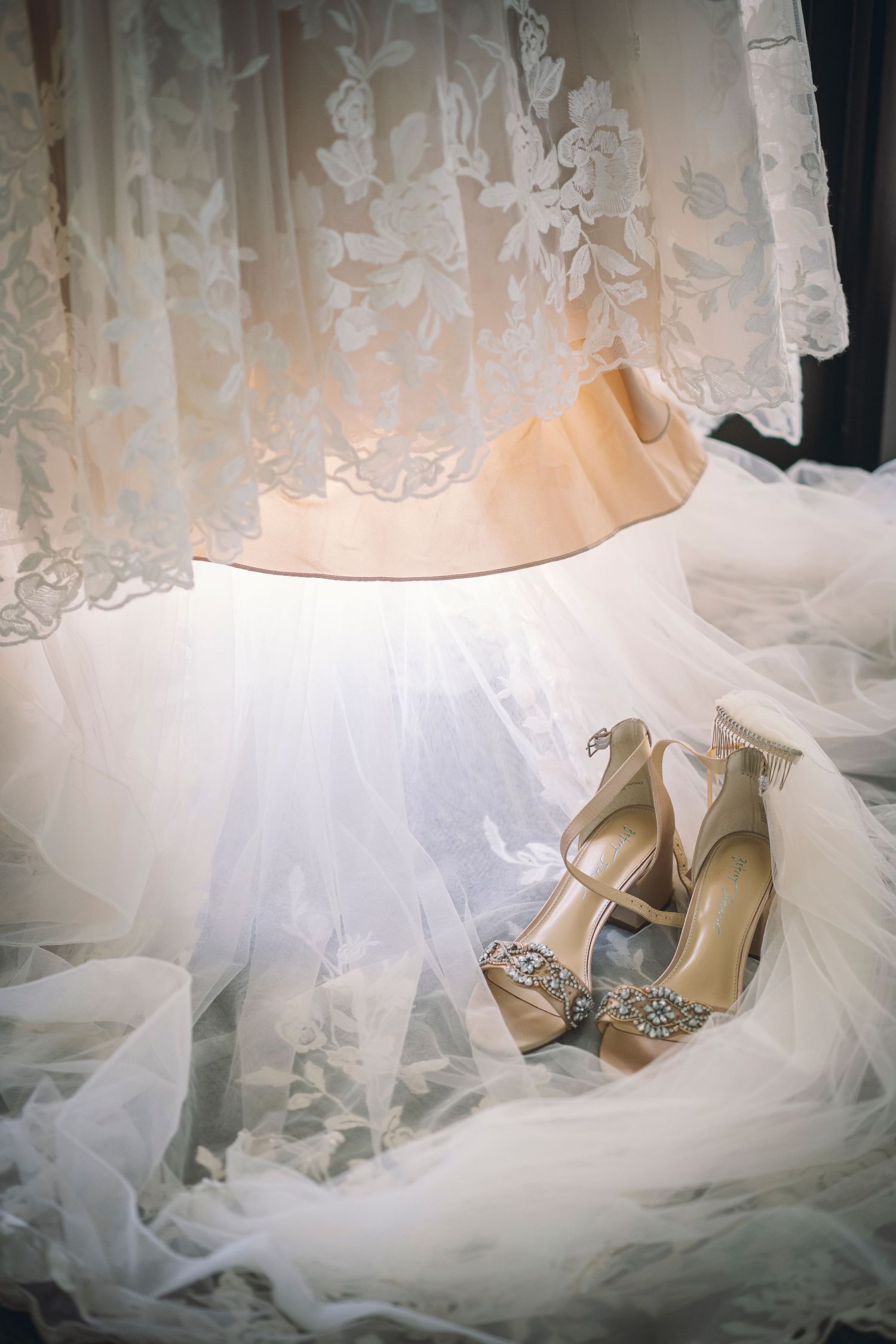 Elegant bridal shoes with crystal embellishments sit against flowing white tulle and lace curtains in soft lighting.