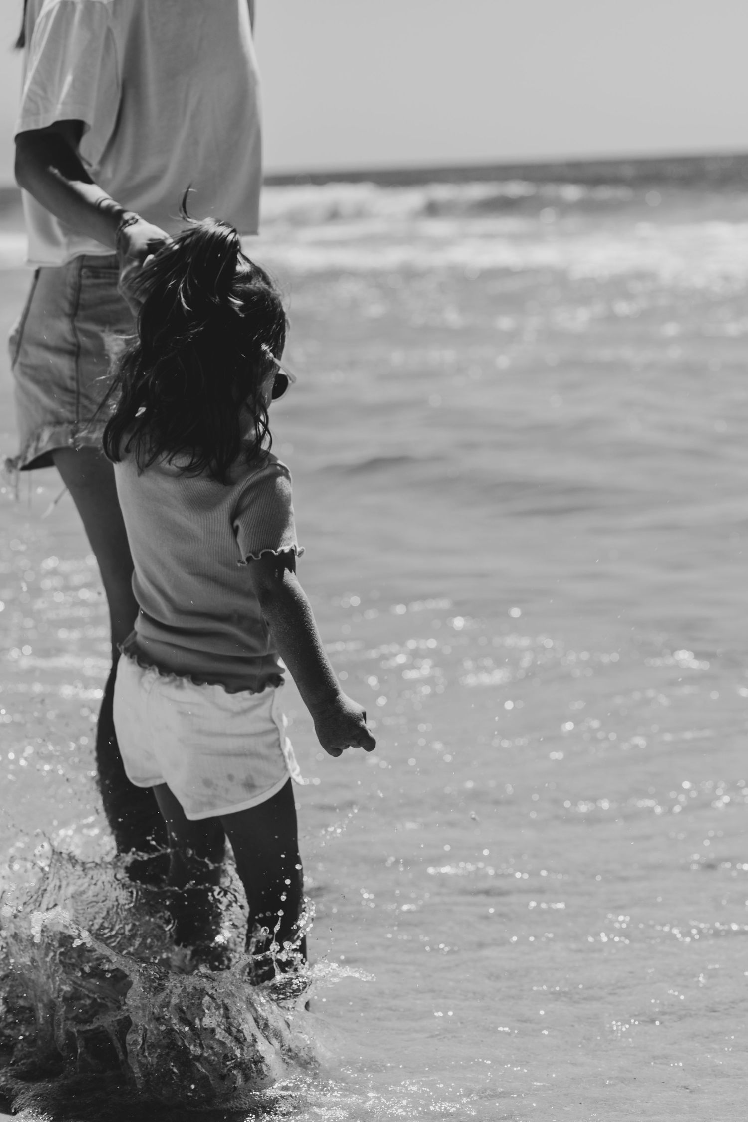A black and white photo of a small child playing in ocean waves while holding an adult's hand.