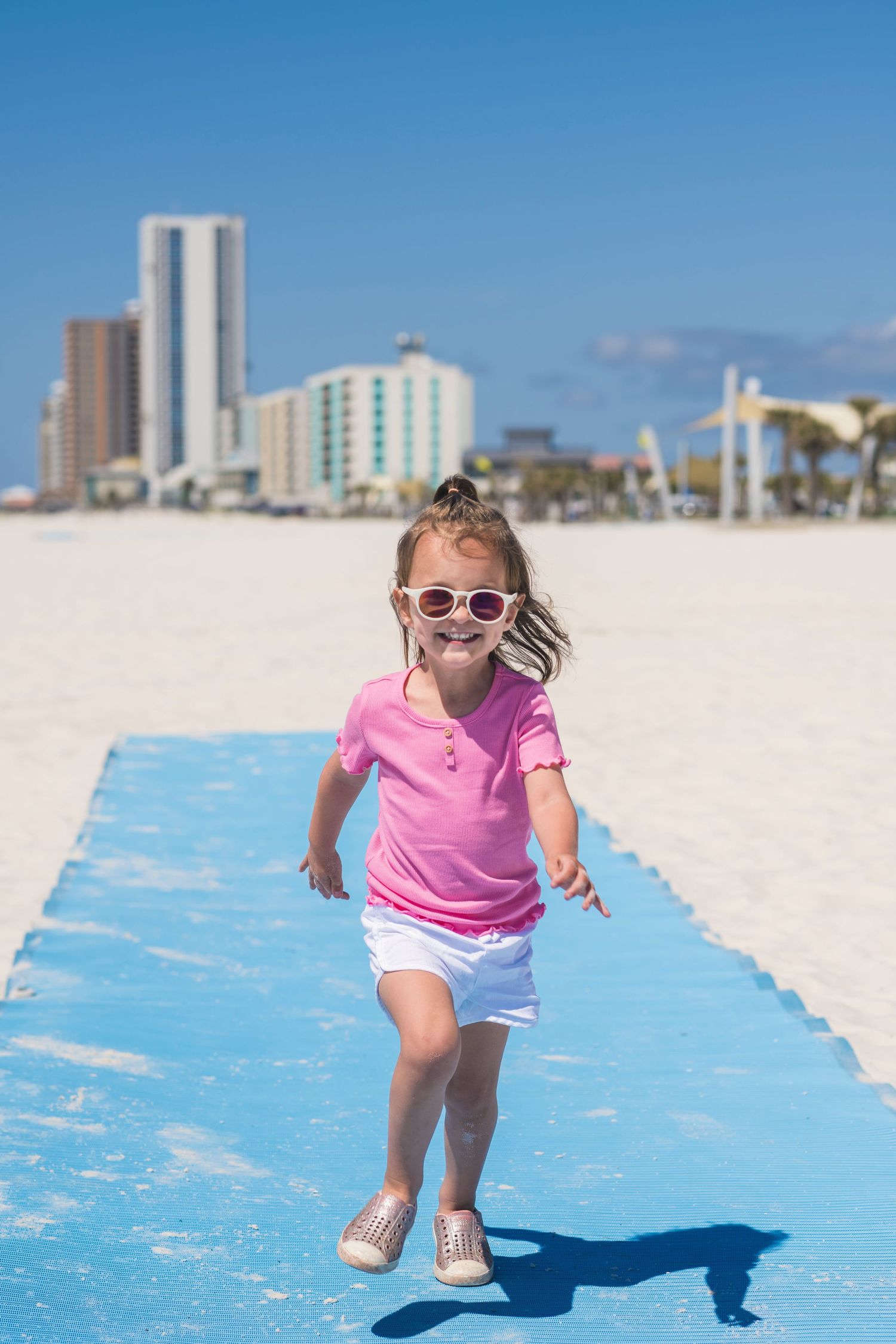 Small child in pink shirt and sunglasses runs along blue boardwalk path on beach with high-rise buildings in background.