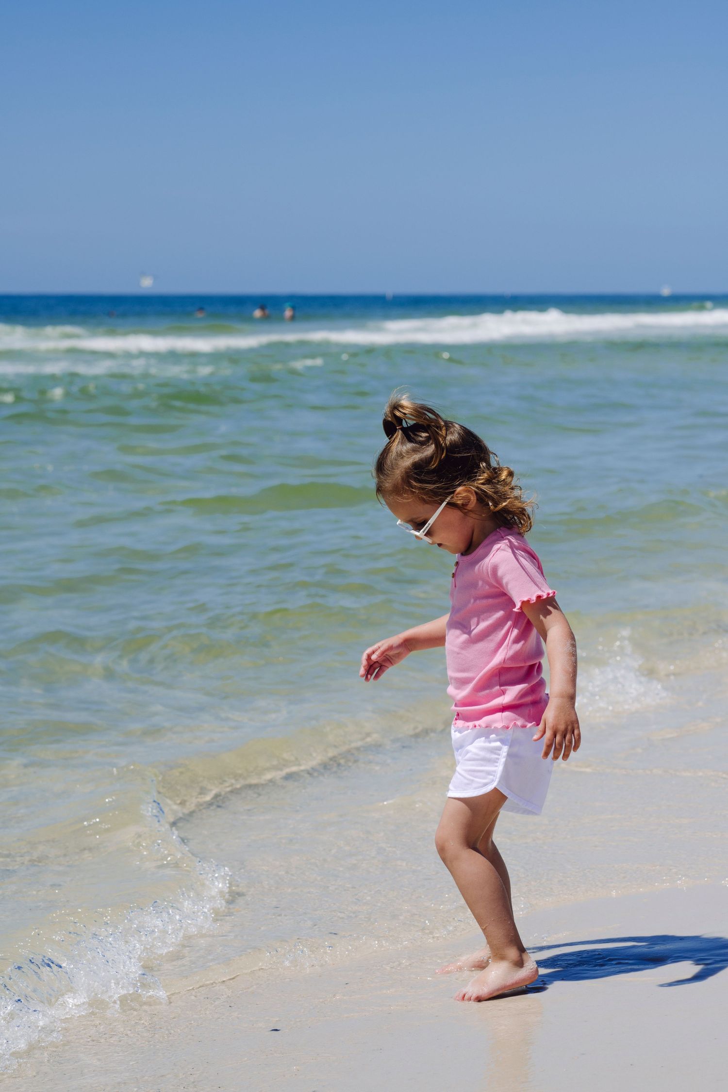 Gulf Shores, AL: Small child wearing pink shirt and white shorts plays along the shoreline at a sunny beach with turquoise waters.