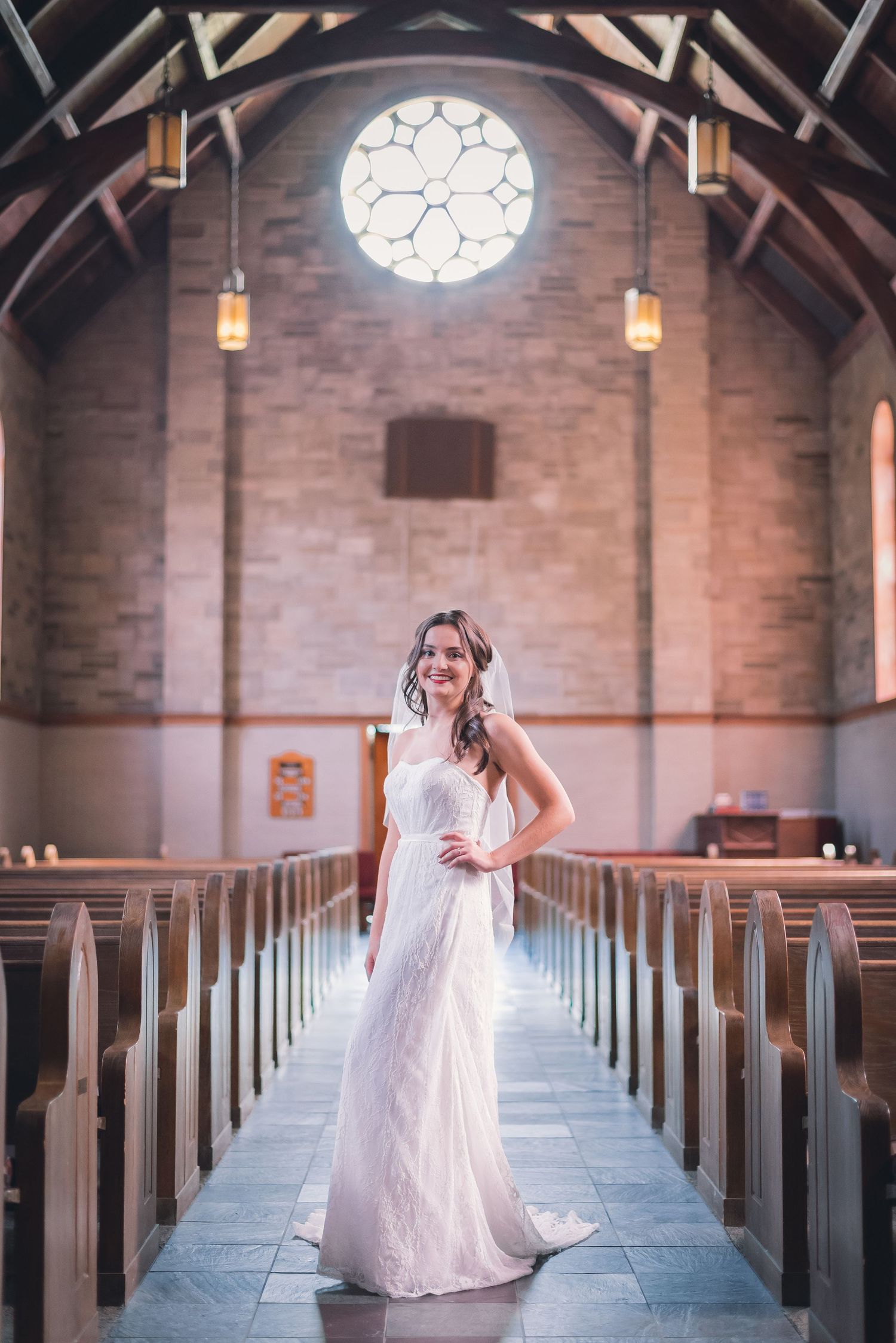 Bridal Portrait with bride posed with natural sunlight creating an elegant scene inside a church's aisle with pews and a rose window.
