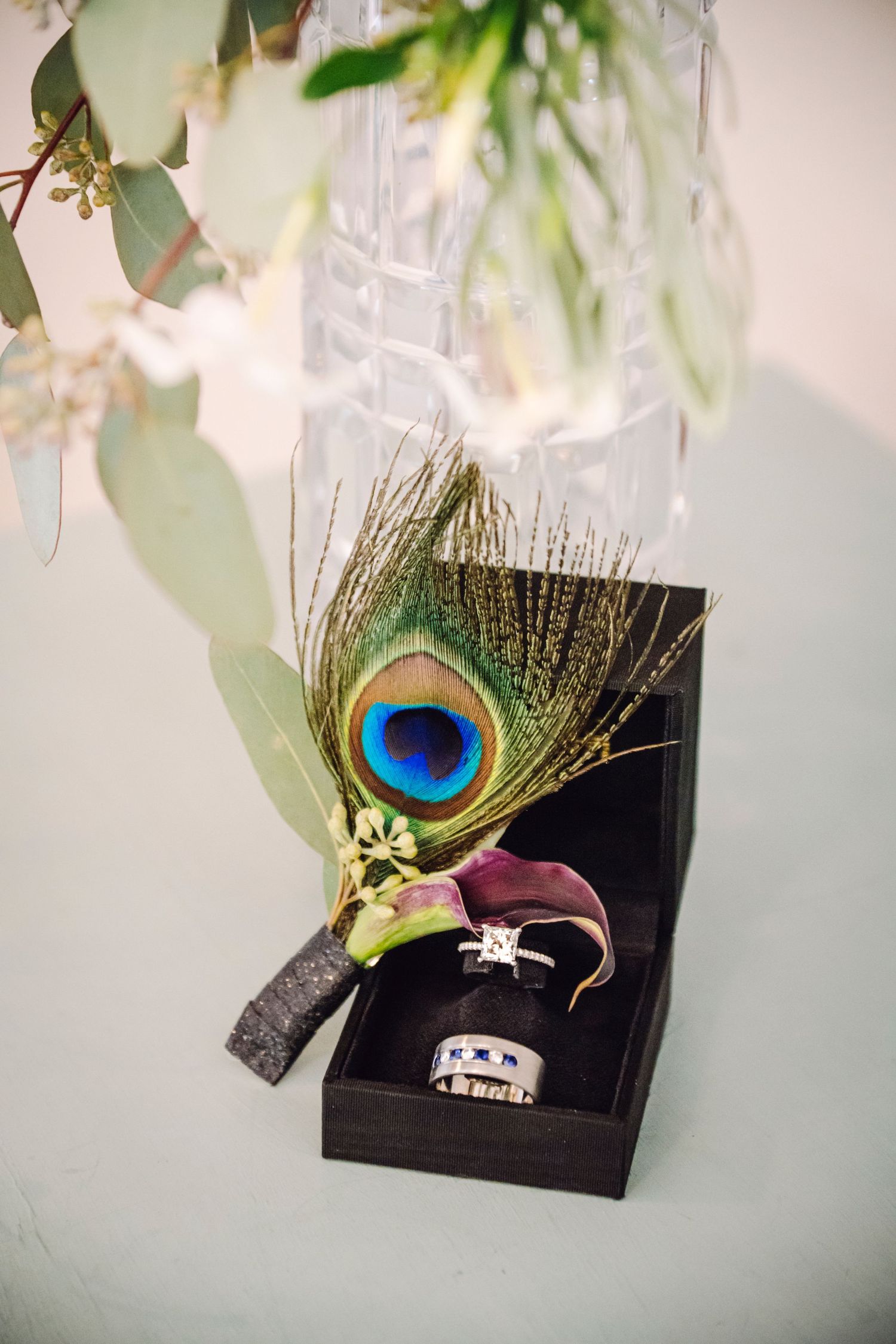 Wedding rings displayed in black box with decorative peacock feather boutonniere accent against white background.