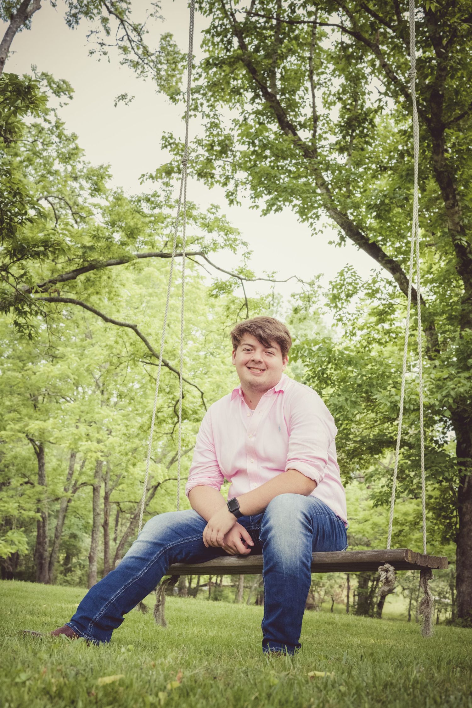 A guy sits on a swing in this Senior Portrait session in a pink shirt and jeans surrounded by a lush green outdoor setting.