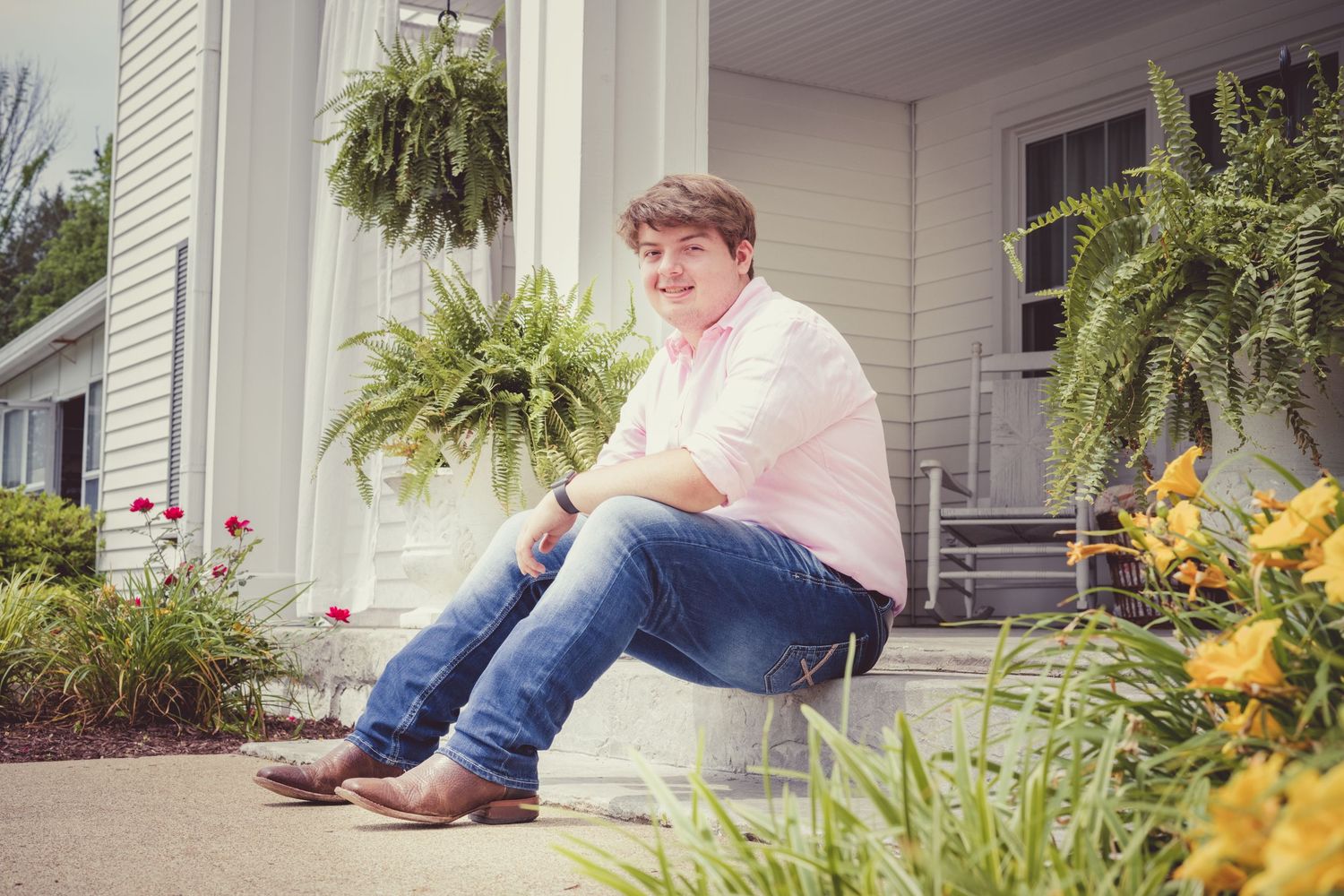 A guy sits casually on a front porch step surrounded by hanging ferns and yellow flowers in this Senior Portrait.