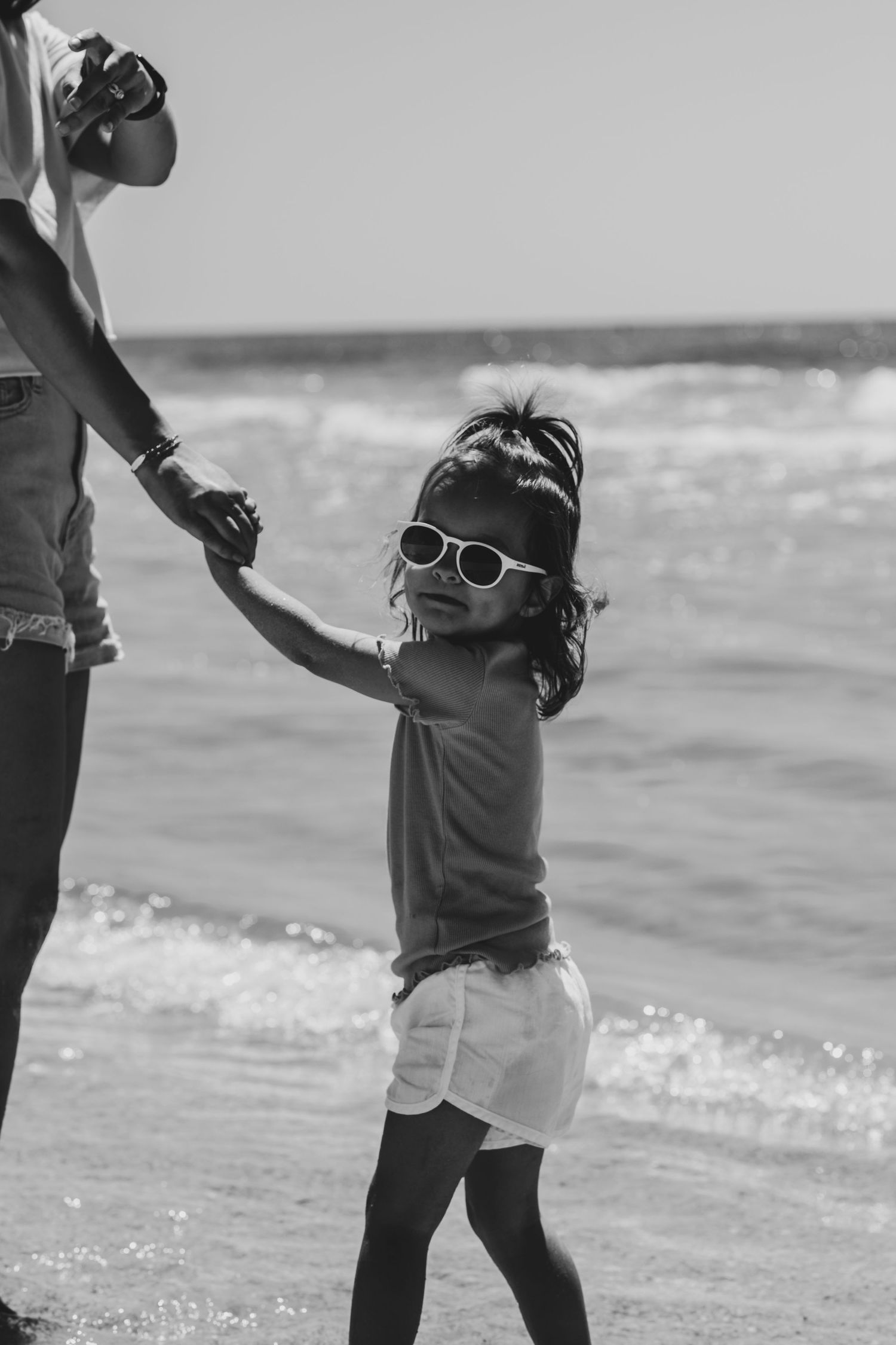 A child in sunglasses holds hands with an adult while enjoying a sunny day at the beach in black and white photography.