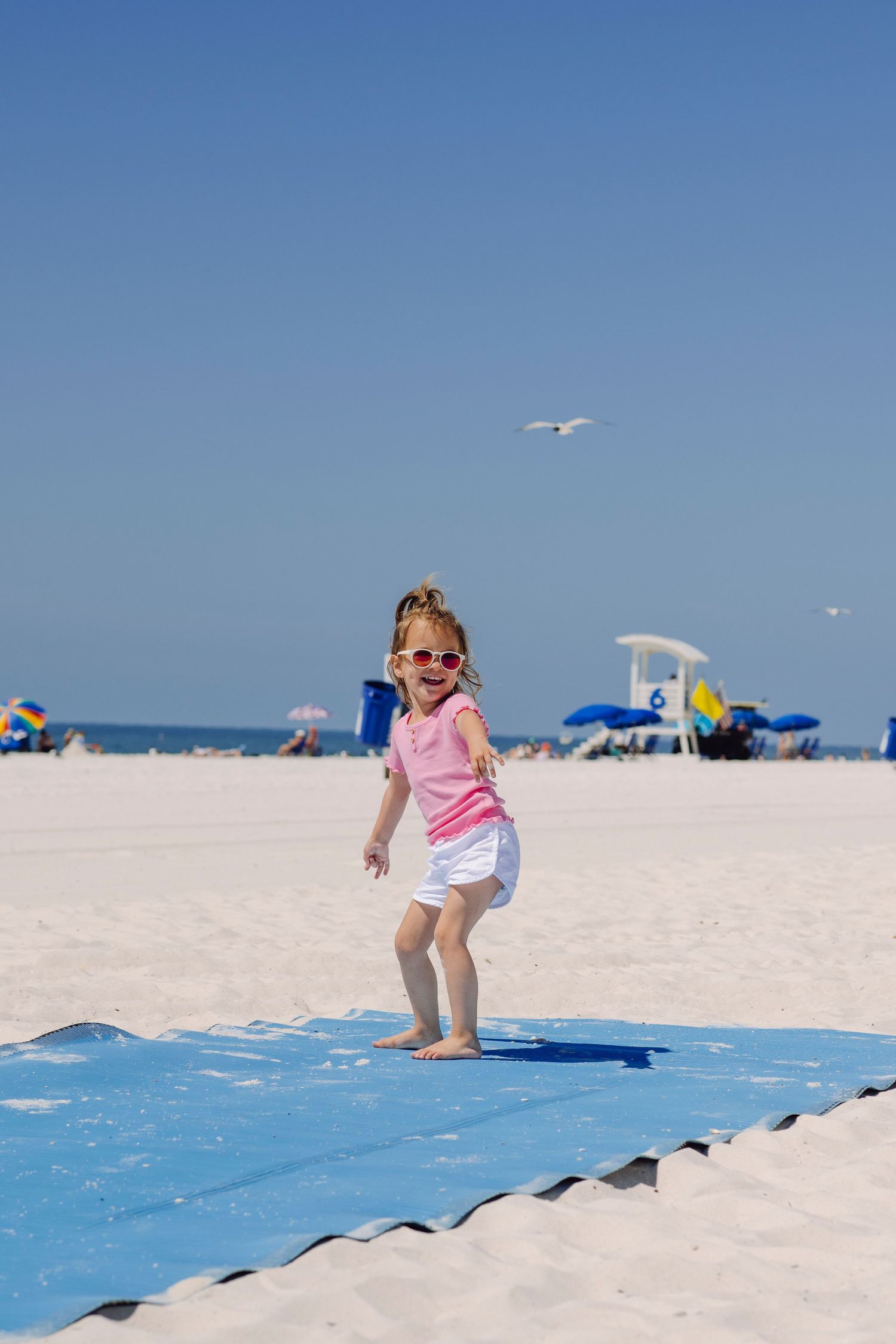 Child playing and jumping on a blue mat at a sunny beach with umbrellas and seagulls in the background.