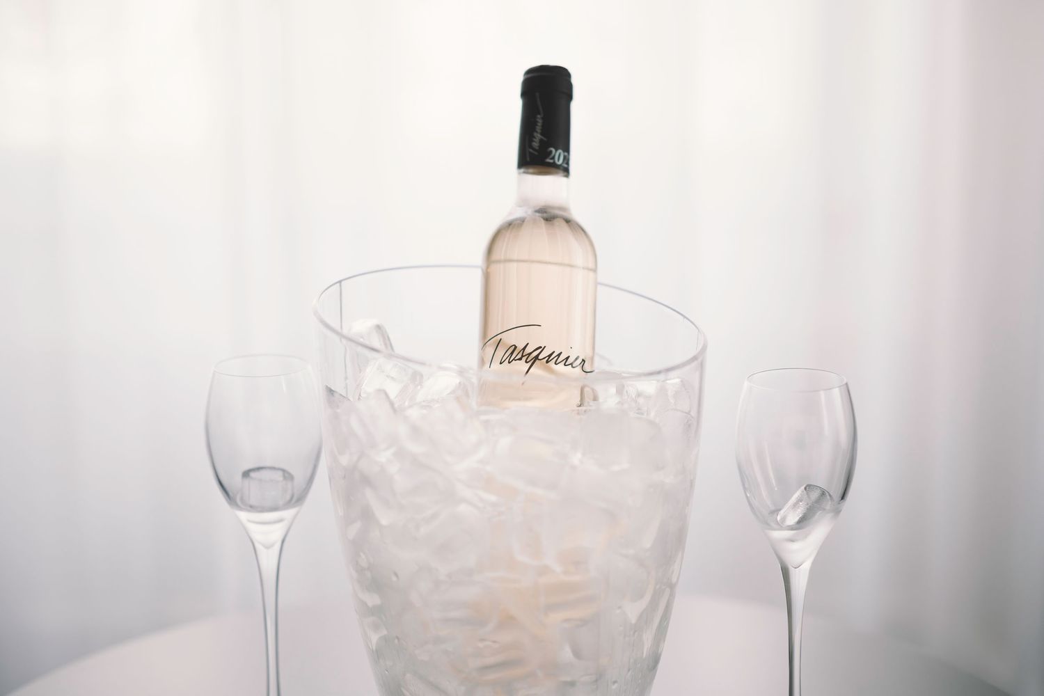 A bottle of white wine chilling in an ice bucket with two empty wine glasses on a white background; steamy French Boudoir session.