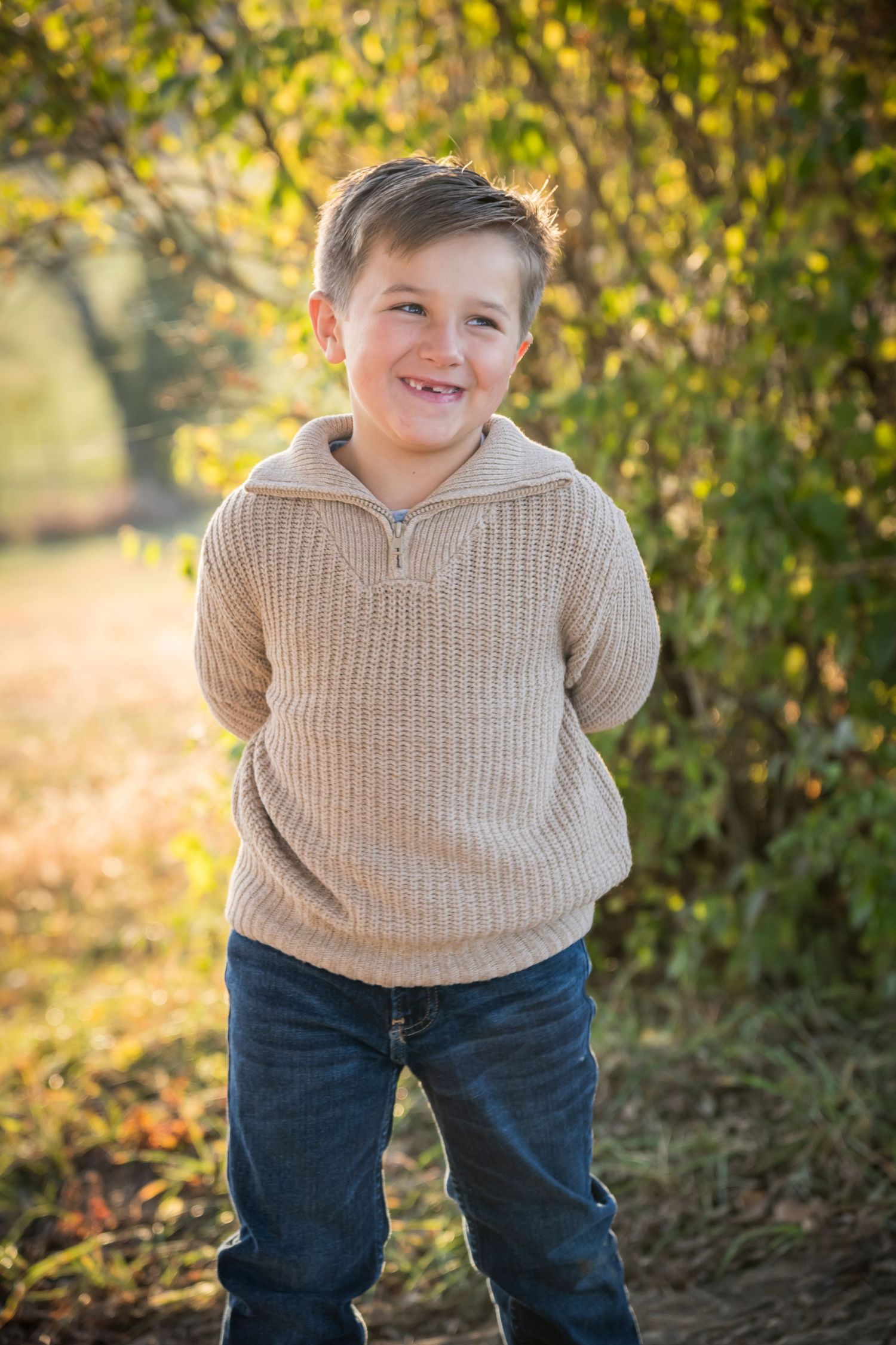 Young child in beige sweater and jeans poses for autumn portrait with golden sunlit foliage in background.