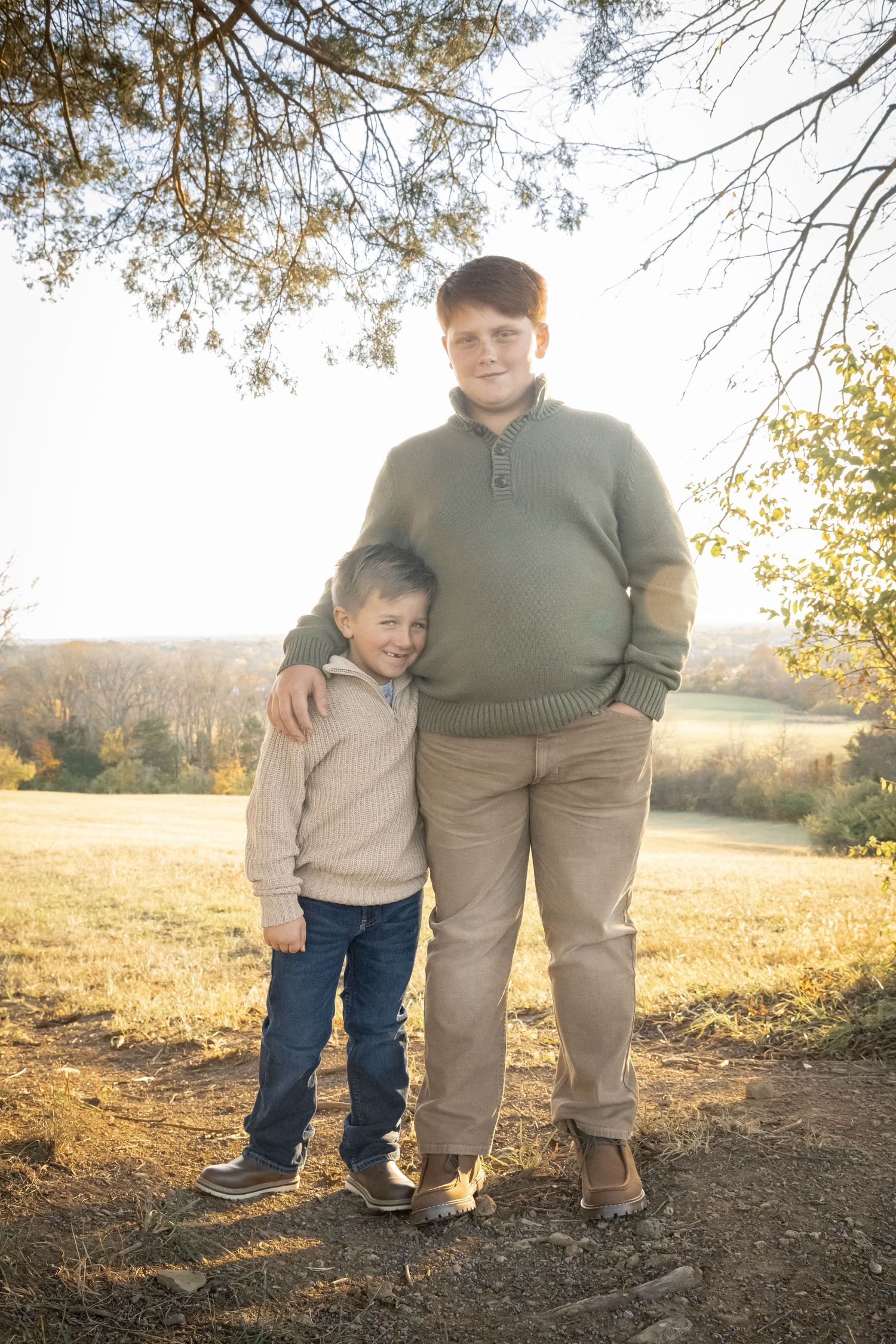 Brothers stand together on a dirt path against a sunlit autumn field and trees during golden hour.
