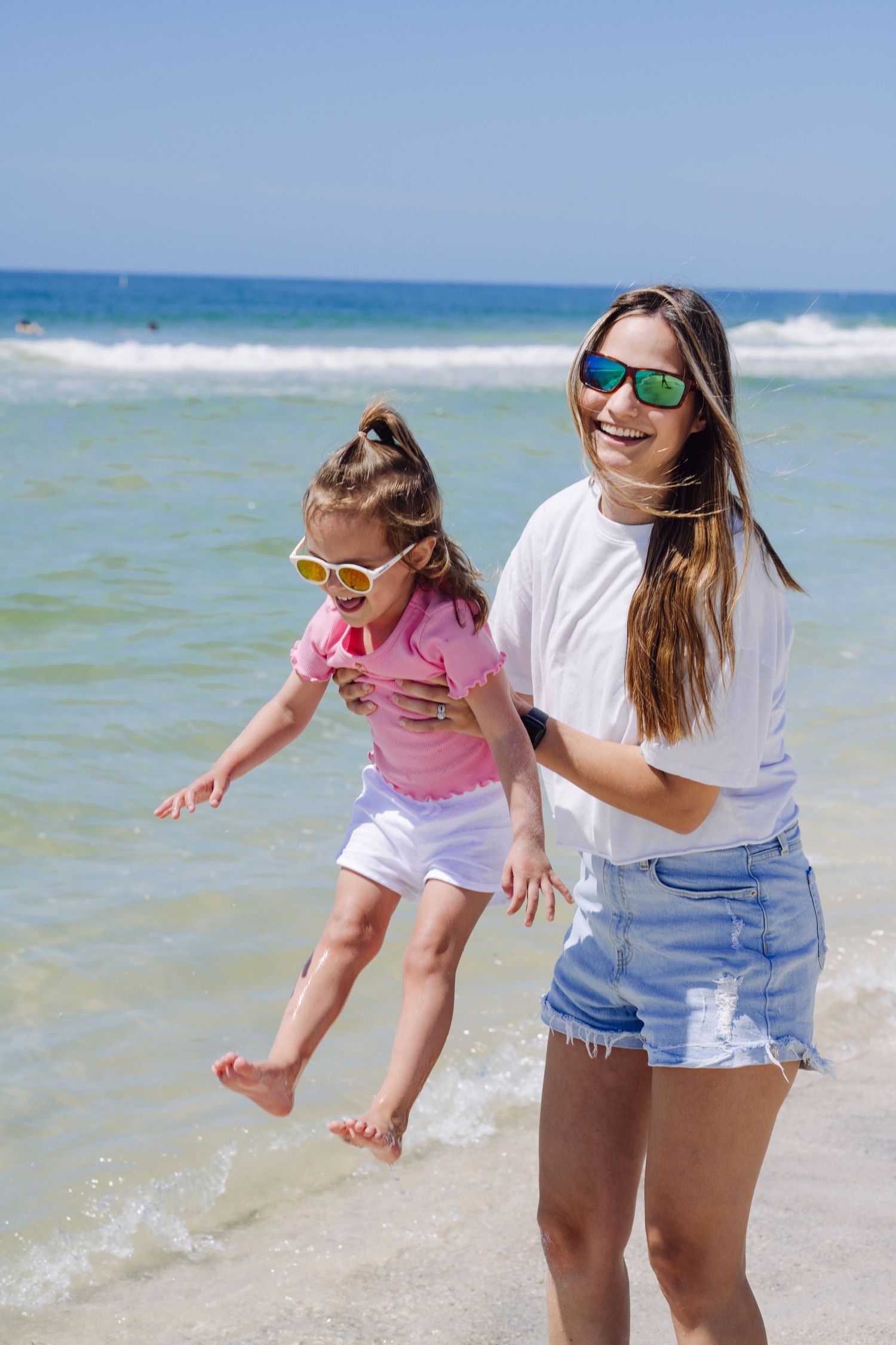Two beachgoers wearing sunglasses enjoy a playful moment in shallow ocean waves on a sunny summer day.