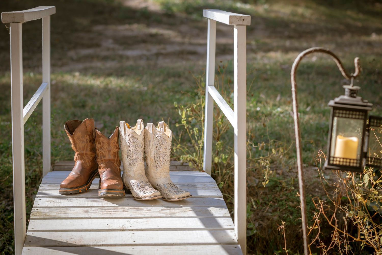 Two pairs of western cowboy boots sit on a wooden platform next to a vintage lantern in a rustic outdoor setting.