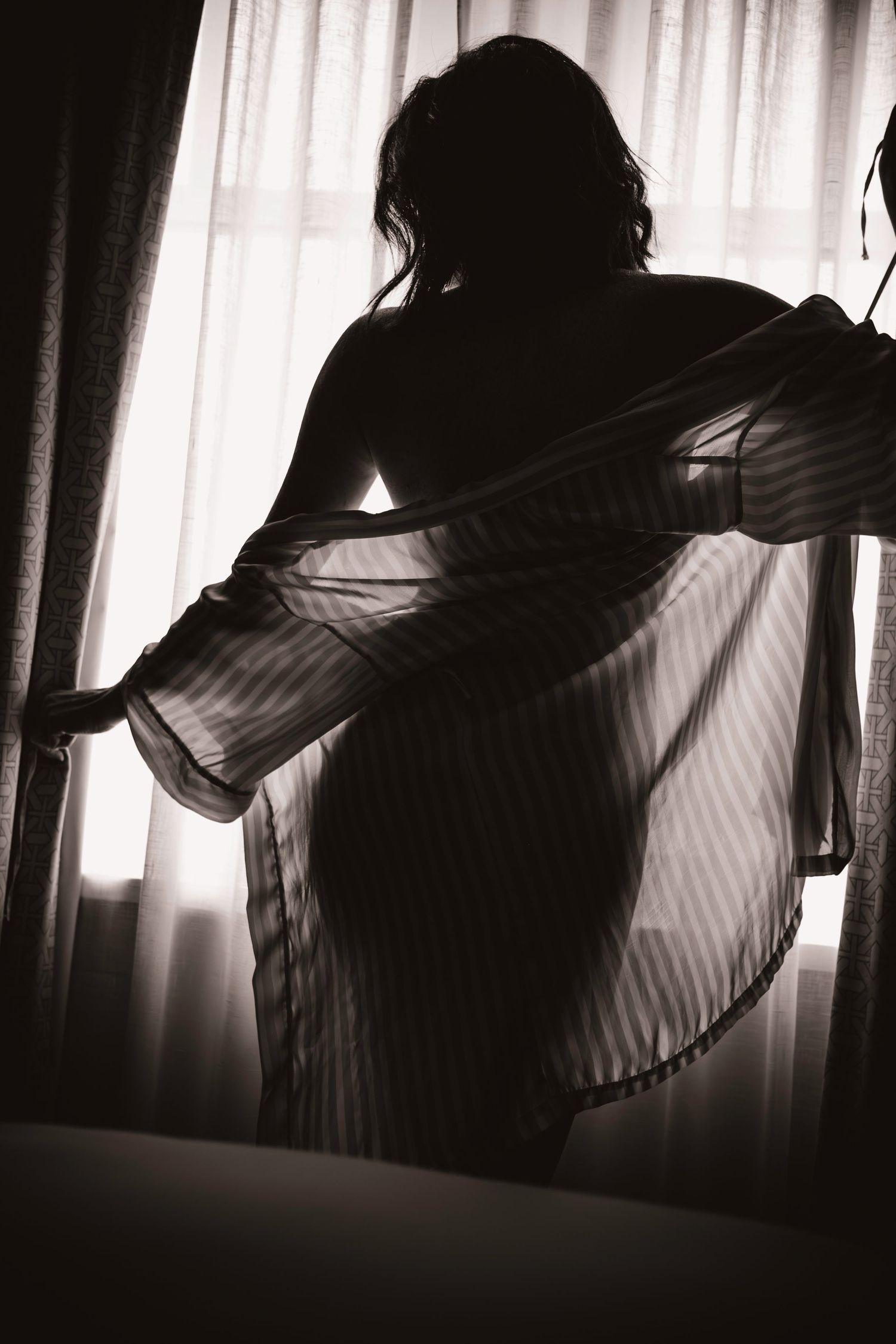 Birmingham, AL boudoir session: silhouetted woman in sheer robe near bright window curtains creates dramatic black and white artistic photo.