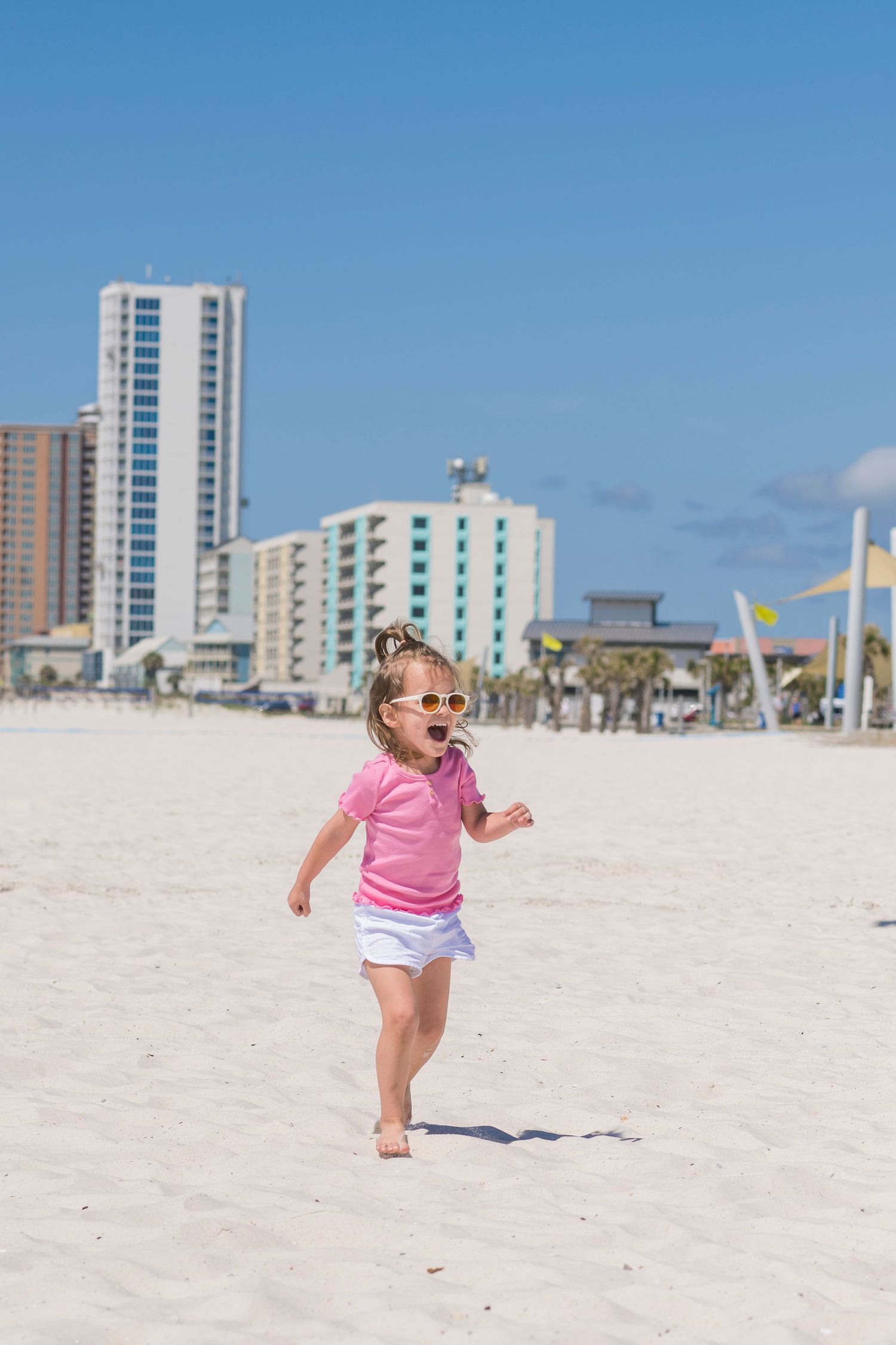 A young child runs joyfully on a white sandy beach with beachfront hotels and condominiums in the background.