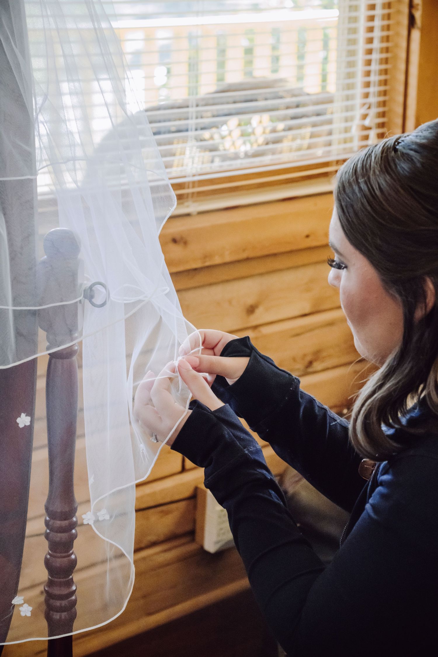 Someone adjusts a delicate white wedding veil by a window in a wooden-paneled room.