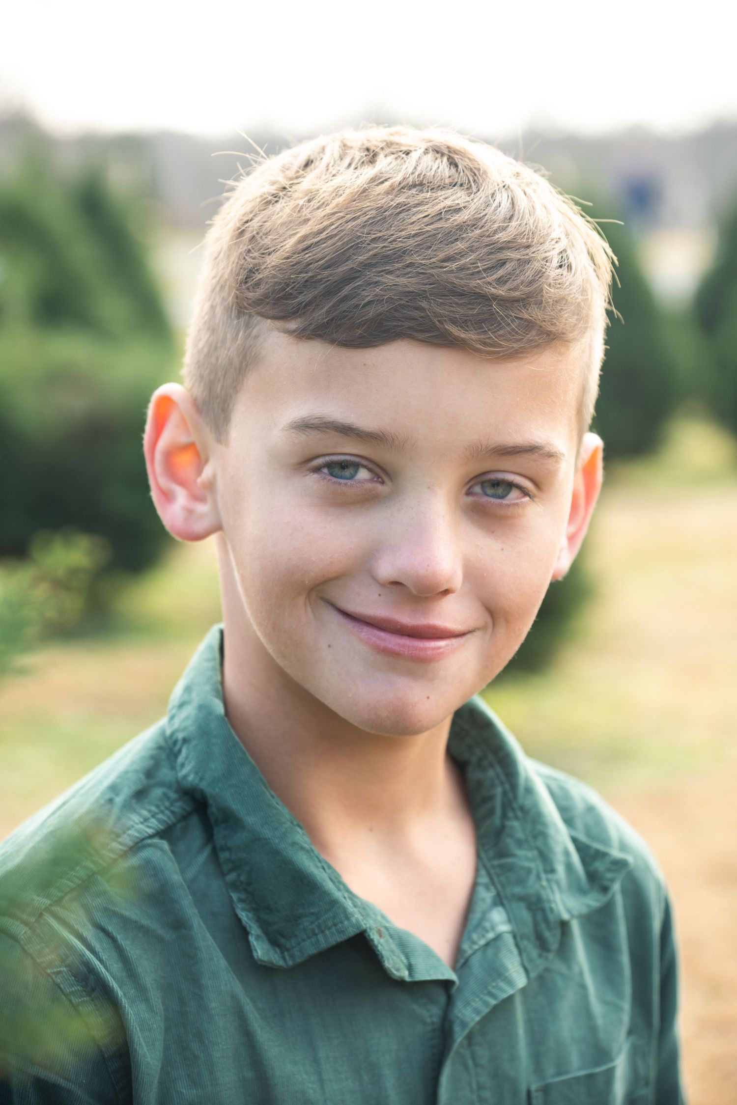 Young boy wearing a green polo shirt posing outdoors on a sunny day in a Christmas tree farm in this Christmas lifestyle session.