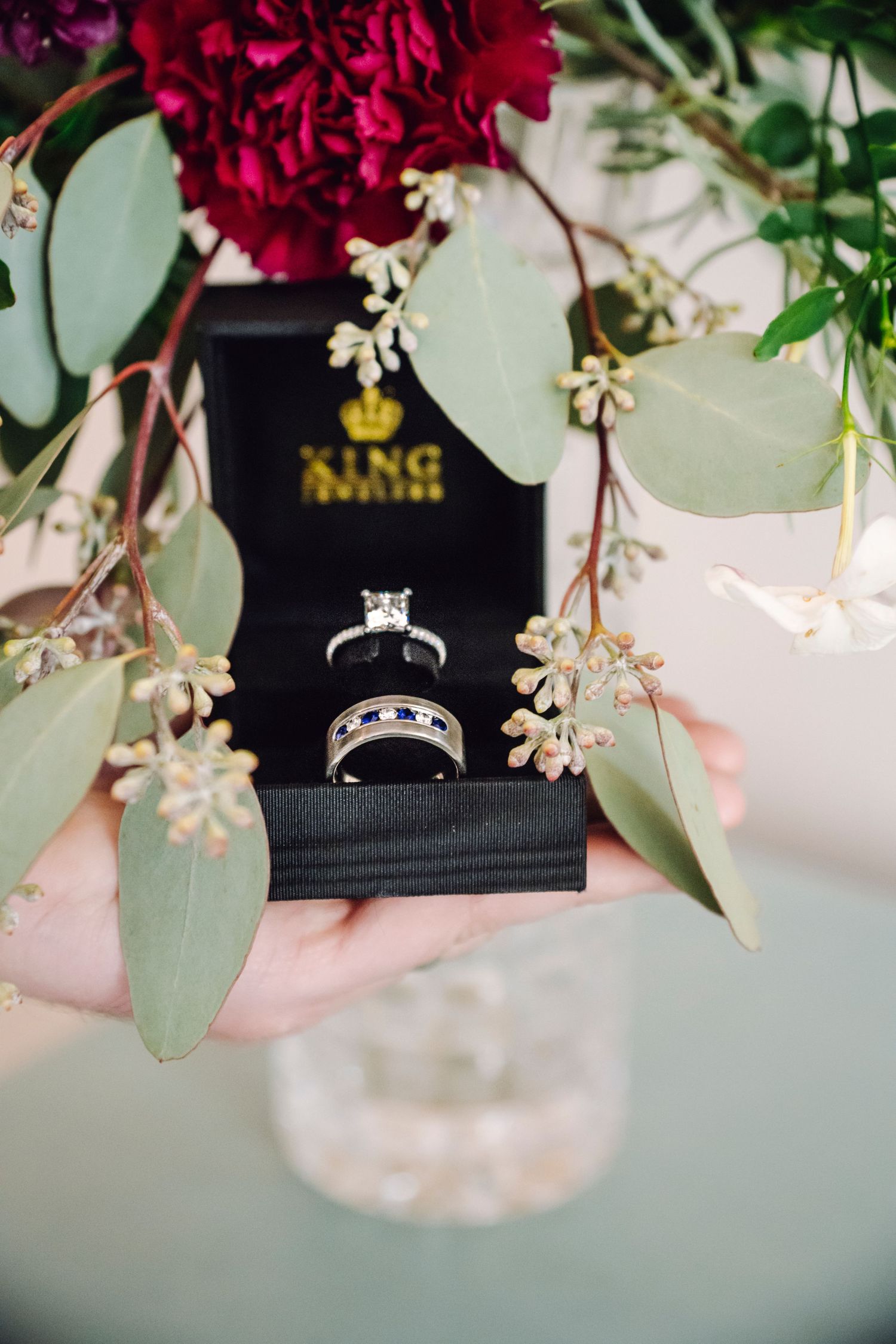 An engagement ring displayed in a black box surrounded by delicate white flowers and burgundy blooms with eucalyptus leaves.