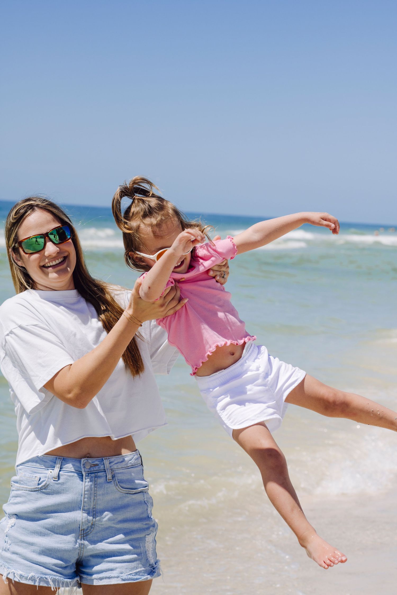 Joyful moment at the beach as parent lifts child wearing pink into the air while waves roll in on a sunny day.