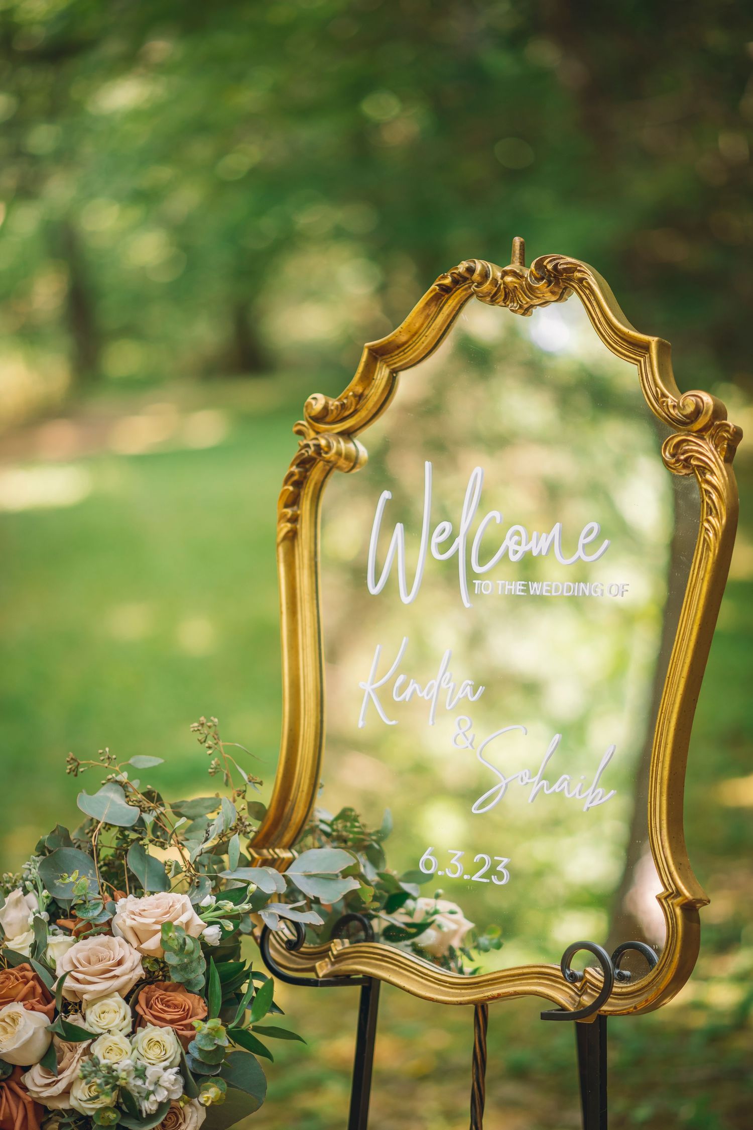 Elegant gold framed mirror signs with white calligraphy lettering and floral decorations displayed outdoors in a garden setting.