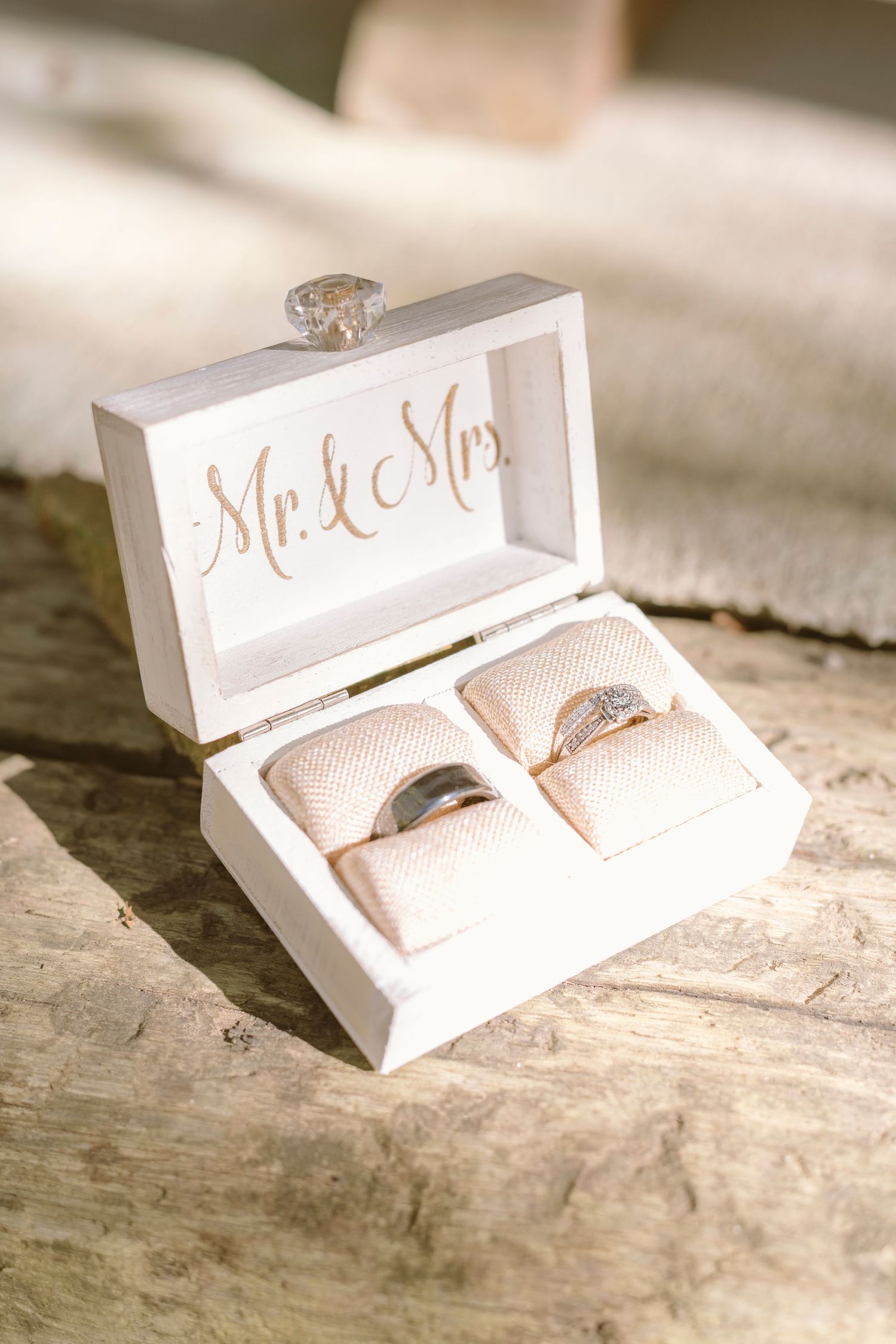 Wedding rings nestled in blush pink cushions inside a white wooden box with 'Mr & Mrs' text on rustic wooden surface.