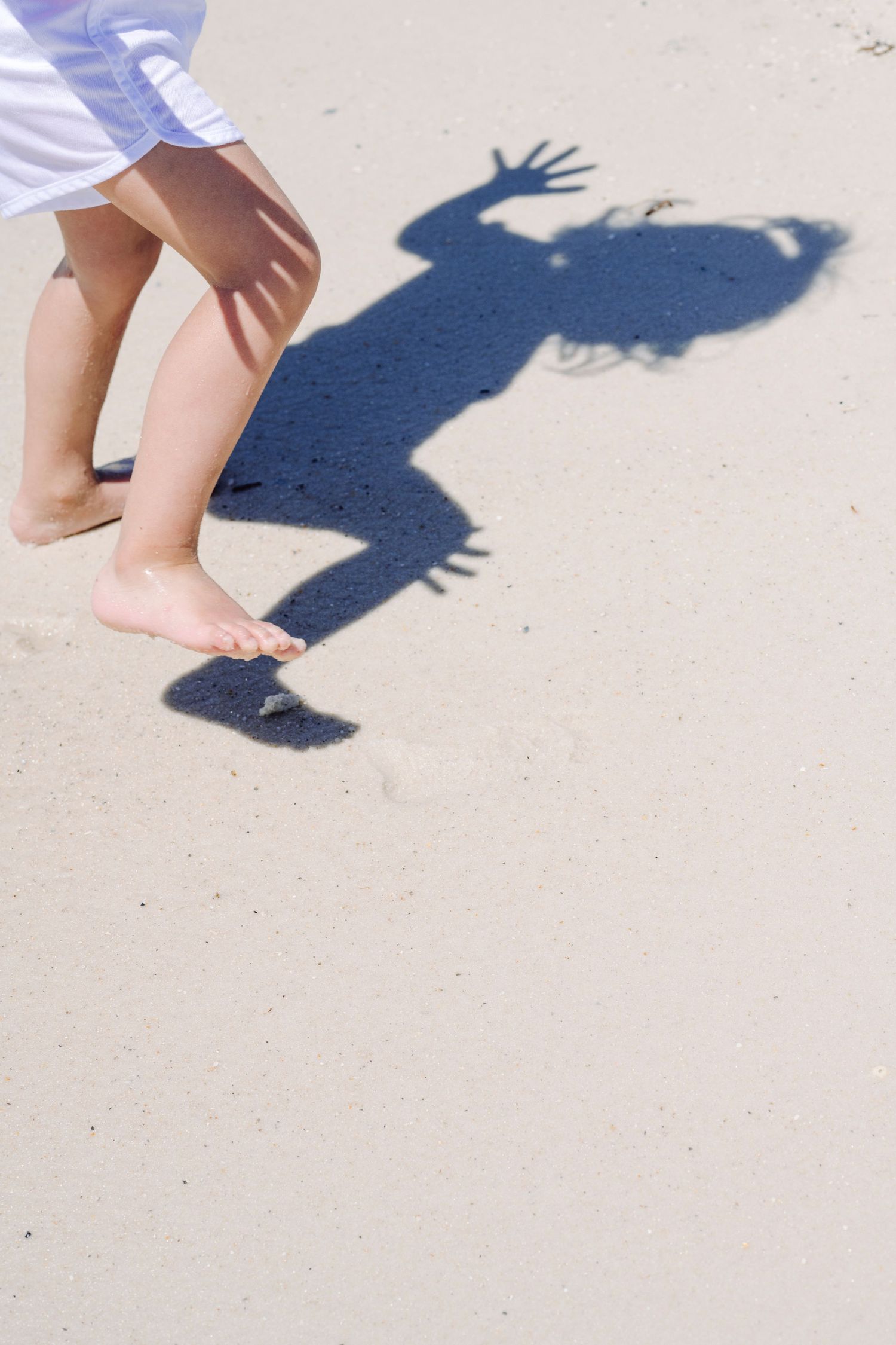 A small child's bare legs and shadow cast on a concrete surface, with the shadow showing raised hands.