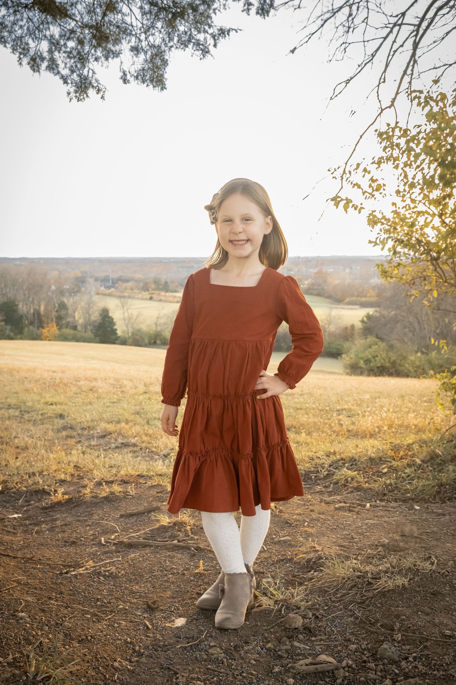 A young girl in a rust-colored tiered dress and white tights stands in a golden autumn field at sunset.